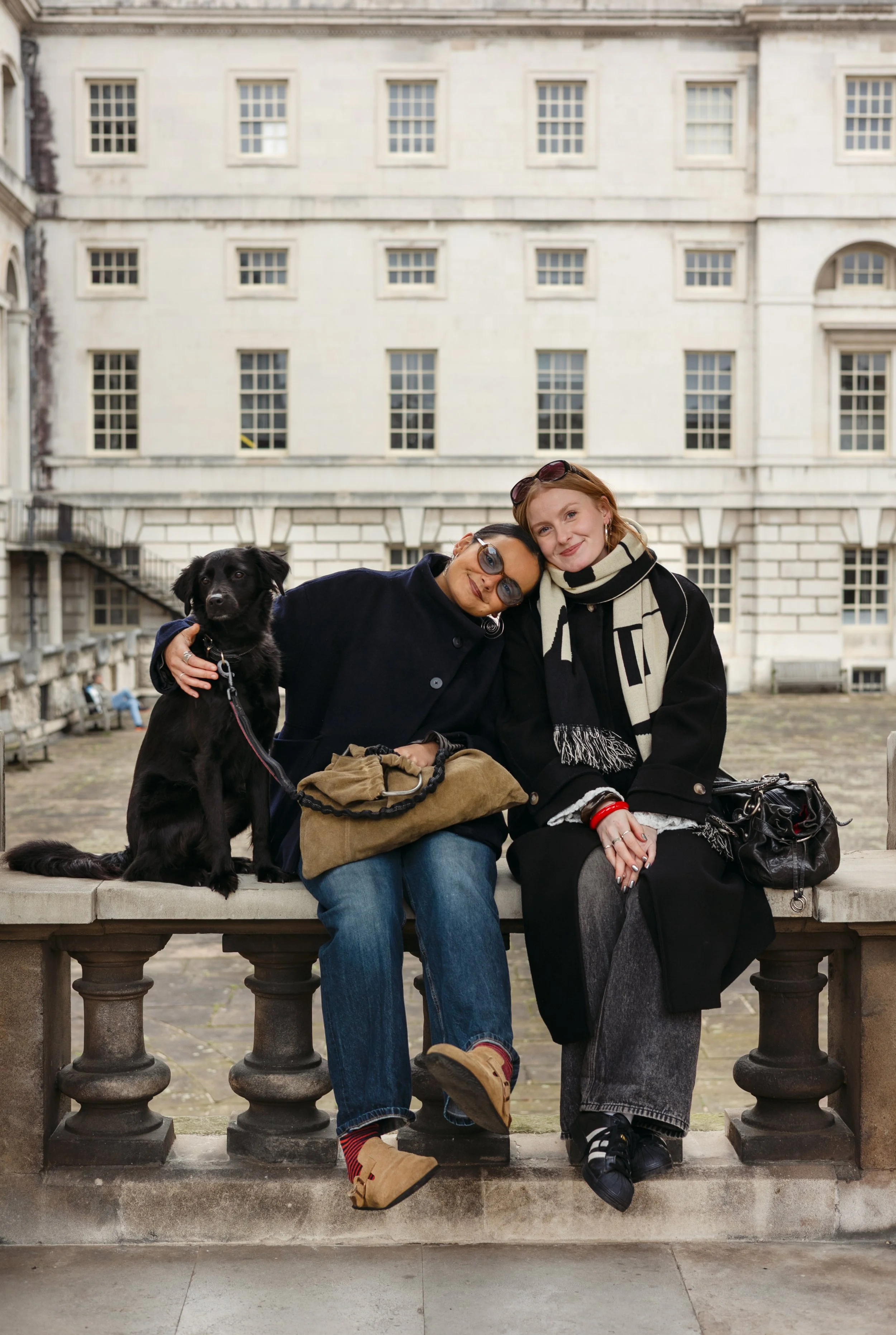 Two women sitting on a stone bench with a black dog. They are outdoors in front of a large cream-colored building with many windows. They are smiling and leaning on each other.