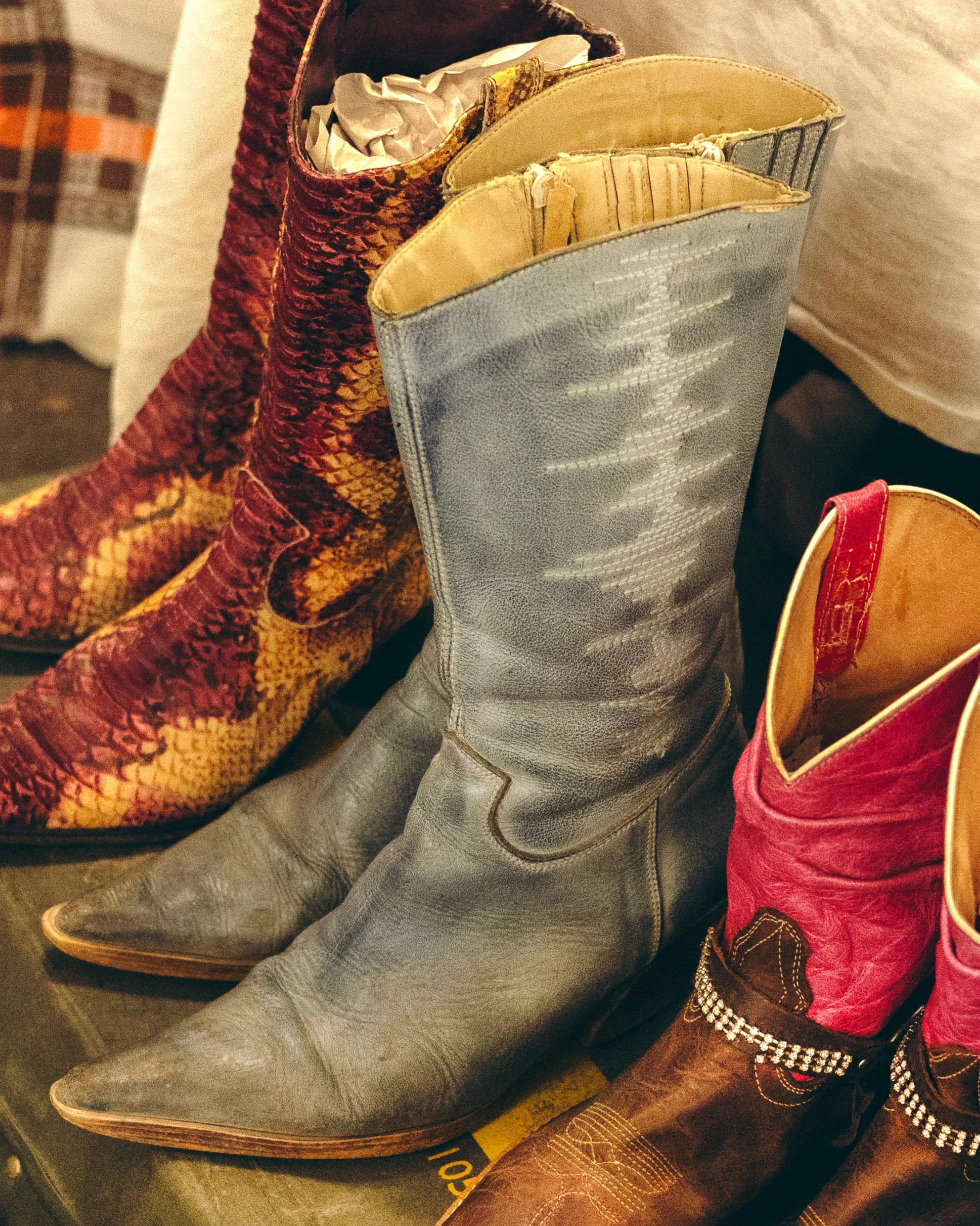 Three pairs of colorful cowboy boots, one with a red and yellow snakeskin pattern, one with a worn gray leather finish, and one with a brown leather base and red upper with decorative straps and rhinestones.