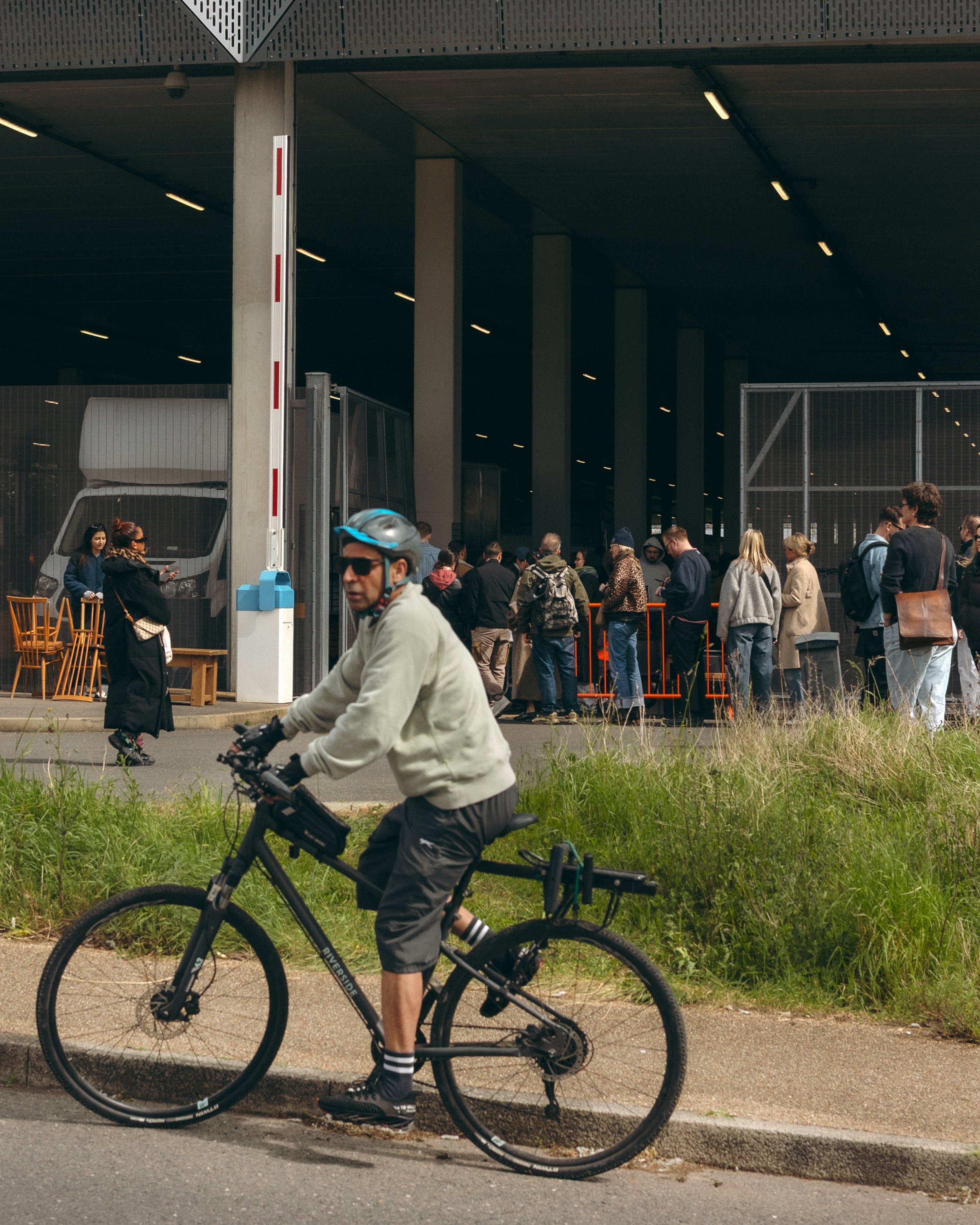A man riding a bicycle in the foreground with a group of people standing in line outside a large building in the background.
