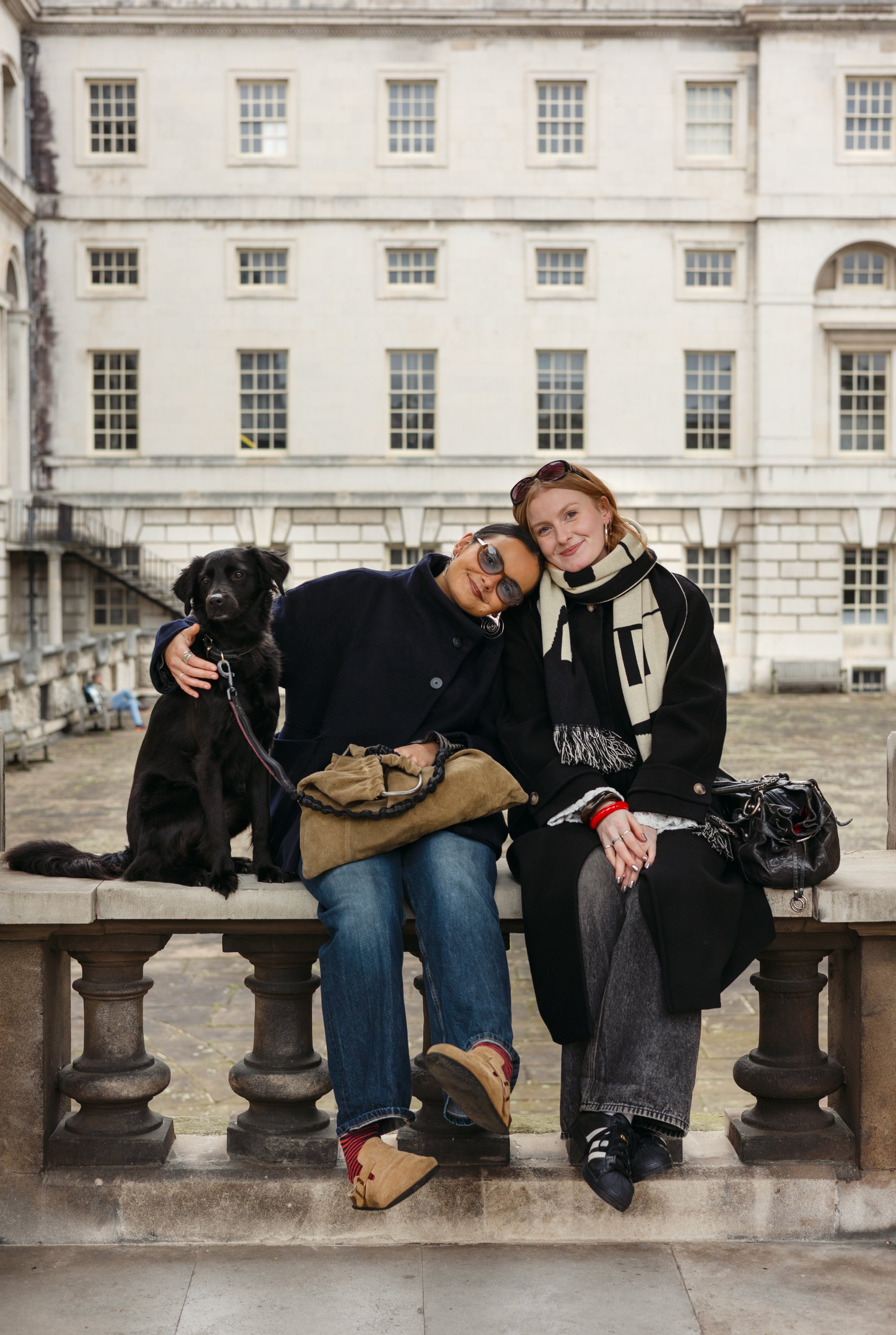 Two women sitting on a stone bench outdoors with a black dog, embracing and smiling. They are wearing casual jackets and scarves. Behind them is a large white building with multiple windows.