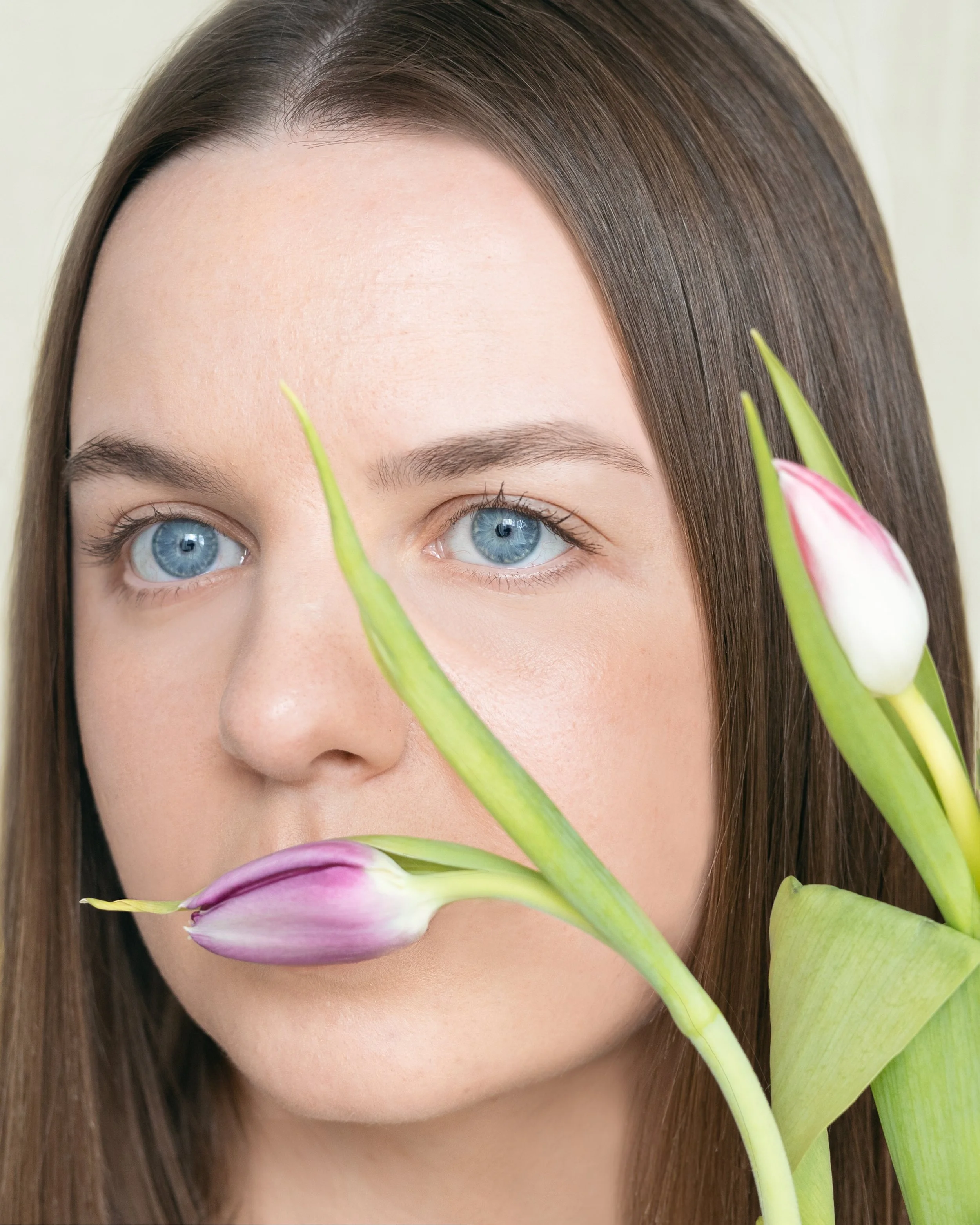 Close-up of a woman's face with blue eyes and brown hair, partially obscured by pink and white tulip flowers in front of her face.