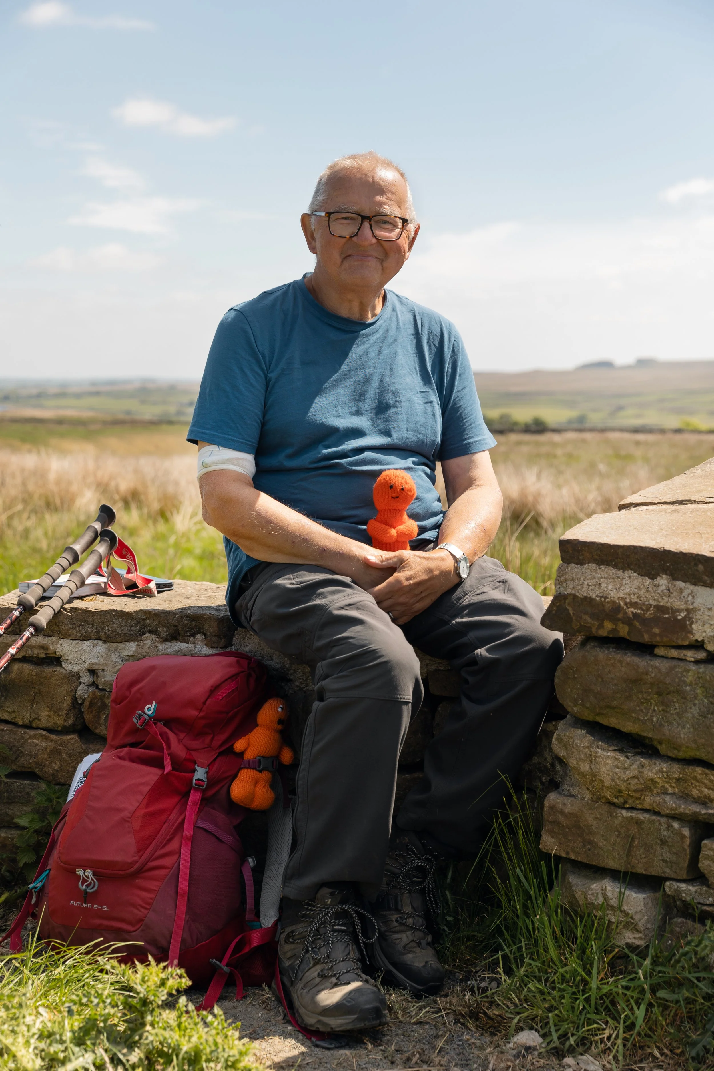 An elderly man sitting on a stone wall outdoors in a grassy field, holding a small orange knitted doll, with hiking poles, a red backpack, and another stuffed doll. He wears a blue t-shirt, gray pants, and hiking boots, and has a bandage on his left 