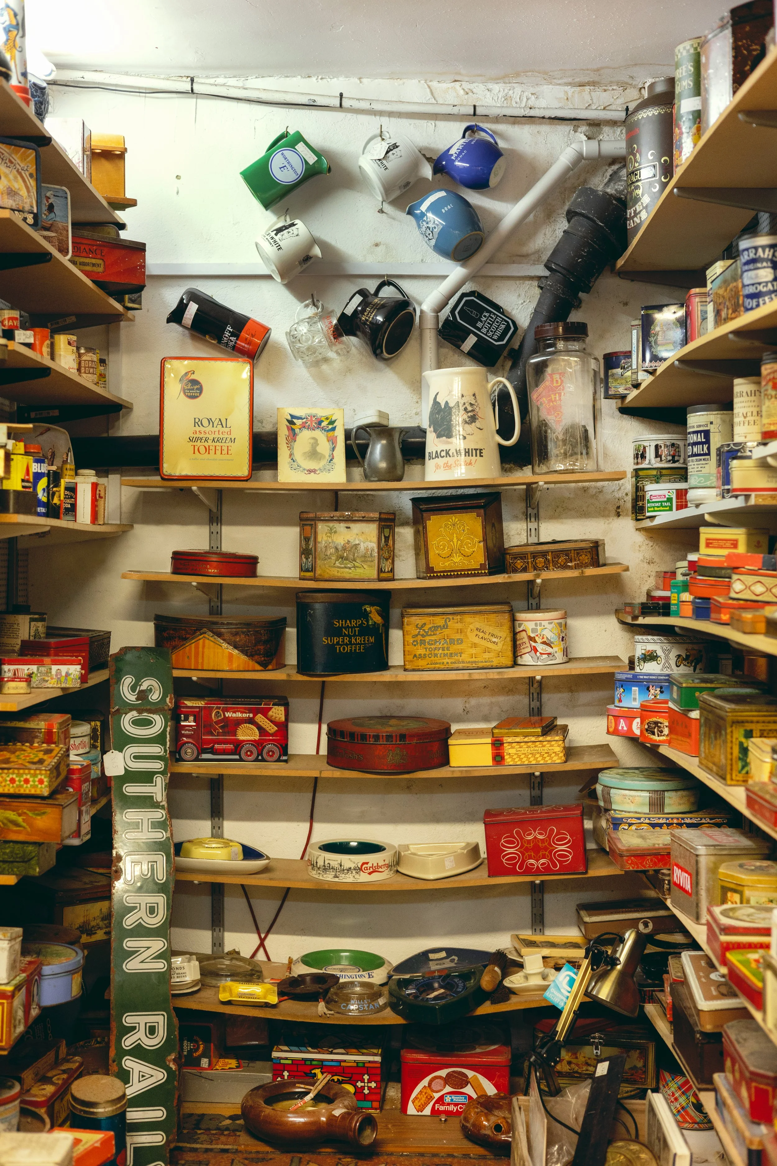 A cluttered storage room with shelves filled with vintage tins, boxes, cans, and household items, including teapots, mugs, and decorative containers.