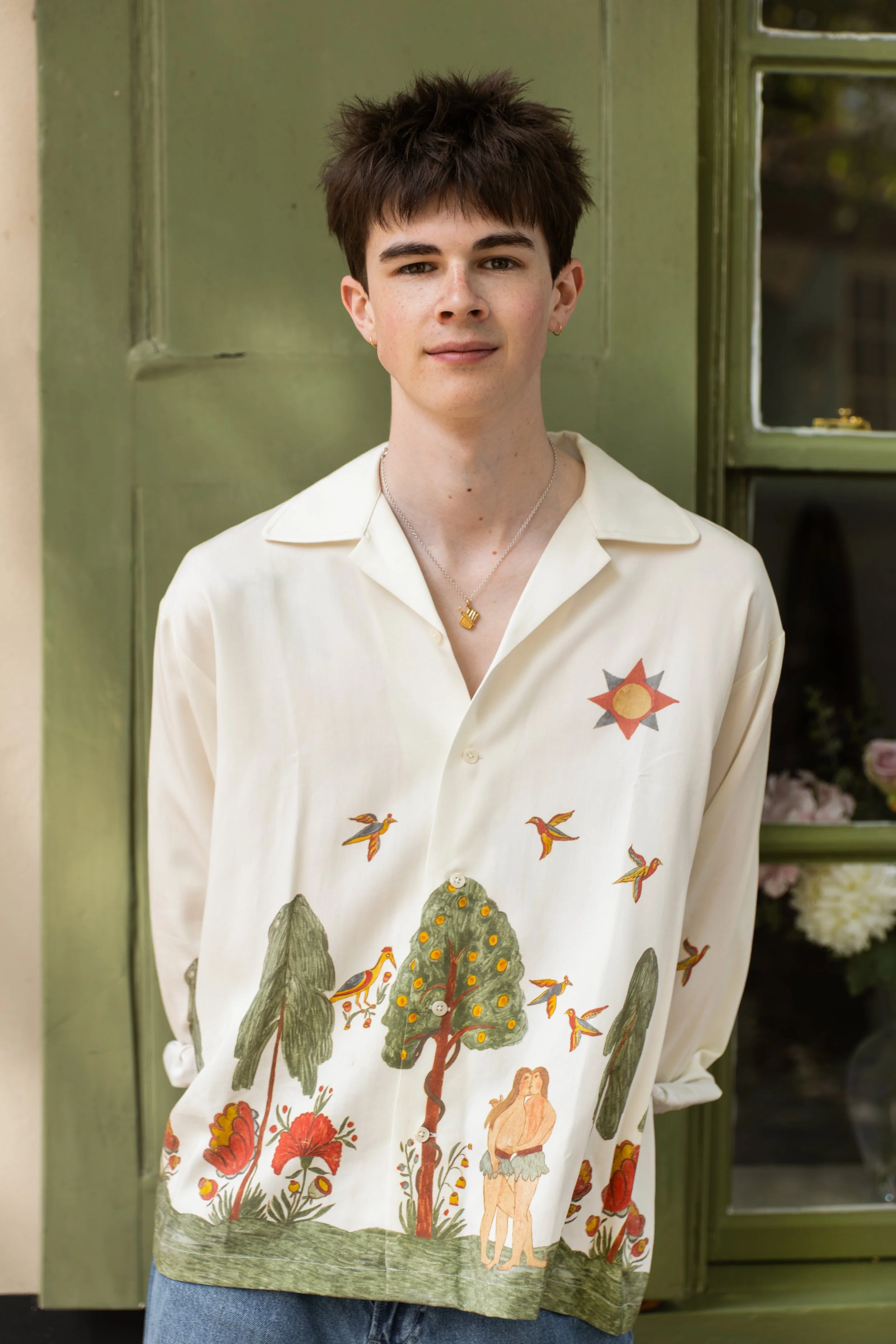 A young person with short, dark, messy hair standing in front of a green door and window, wearing a cream-coloured shirt with colourful nature-themed illustrations of trees, birds, and a necklace with a small pendant.