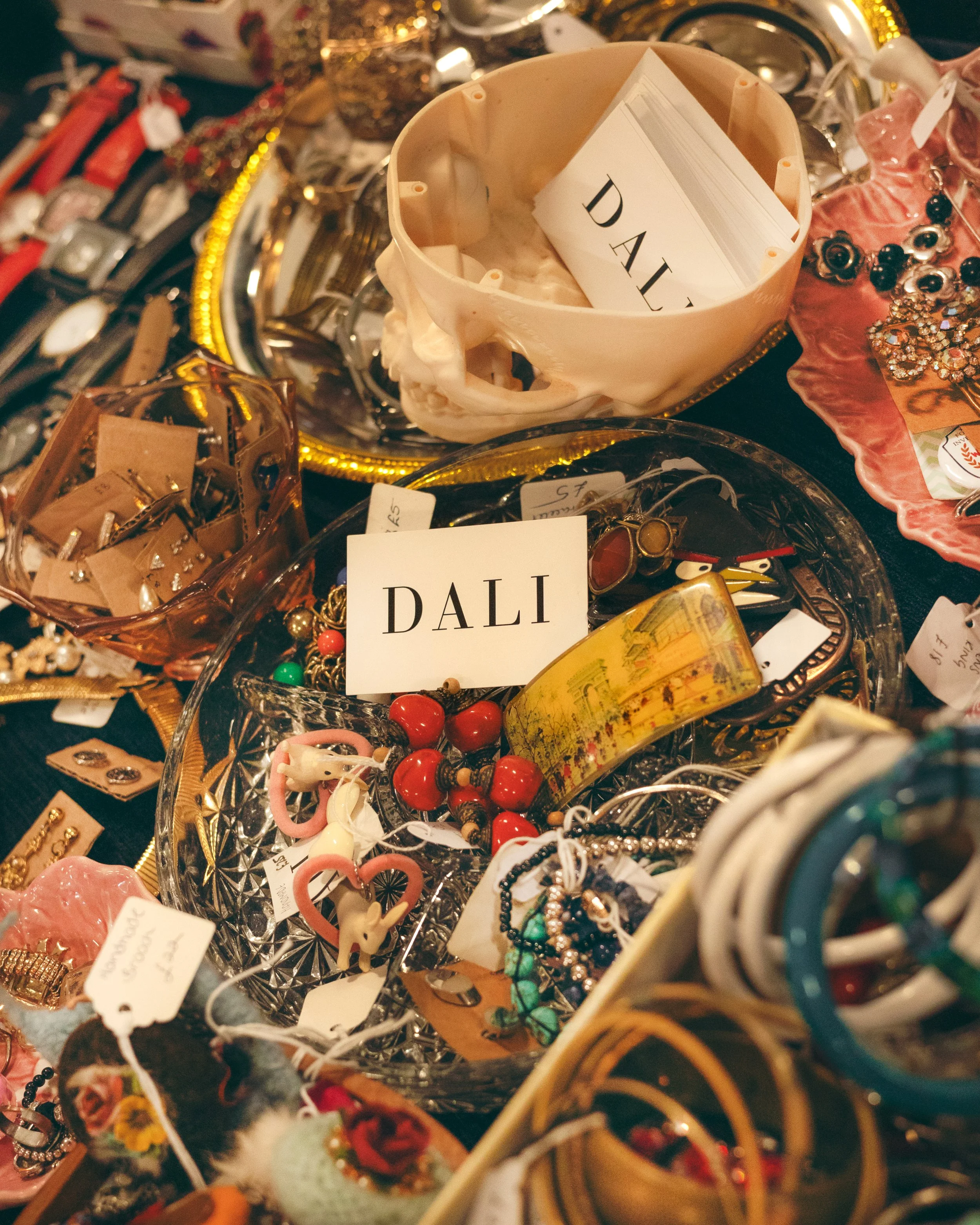 A cluttered table of vintage jewelry and accessories, with bowls and trays holding earrings, necklaces, and bracelets, labeled with the name "DALI."