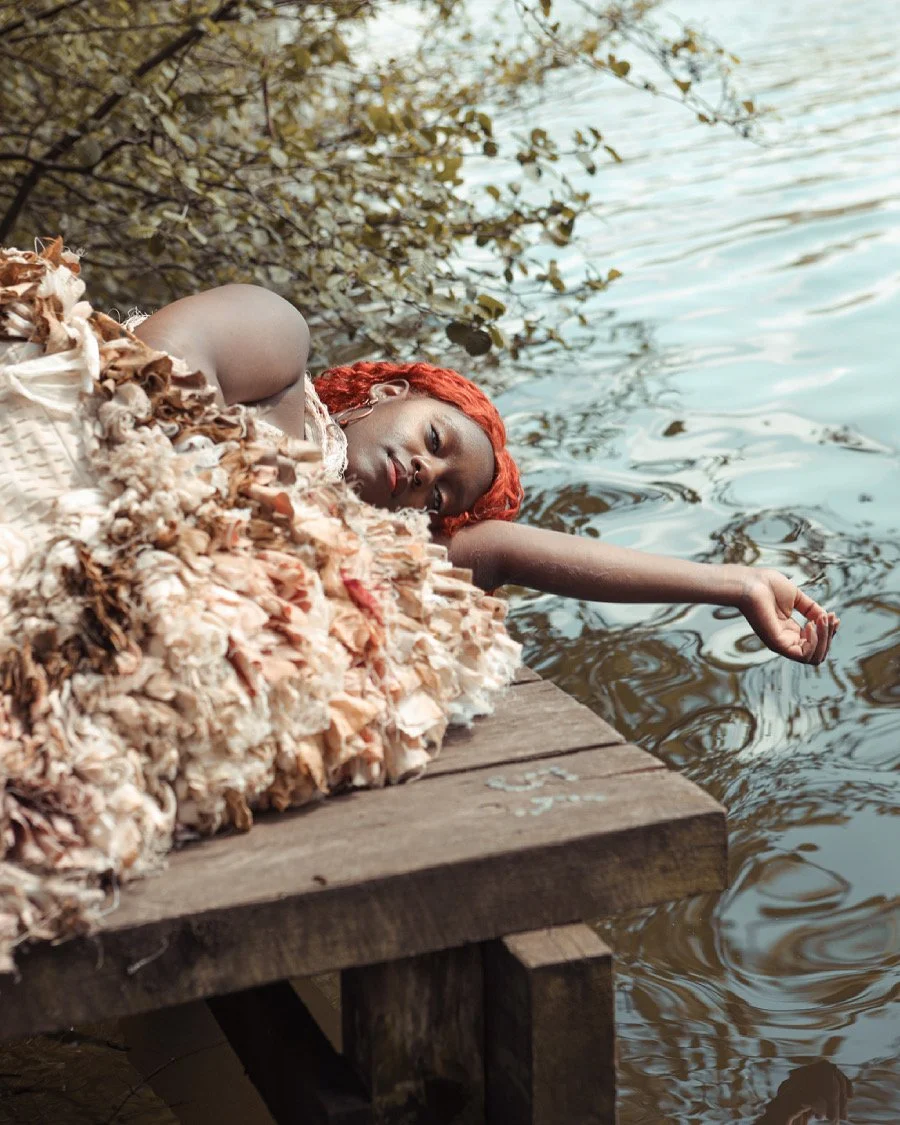 A woman with vibrant red hair laying on a wooden dock by the water, wearing a textured, pastel-colored dress, and extending her arm towards the water. Her eyes are closed, and she appears relaxed, surrounded by trees with overhanging branches.