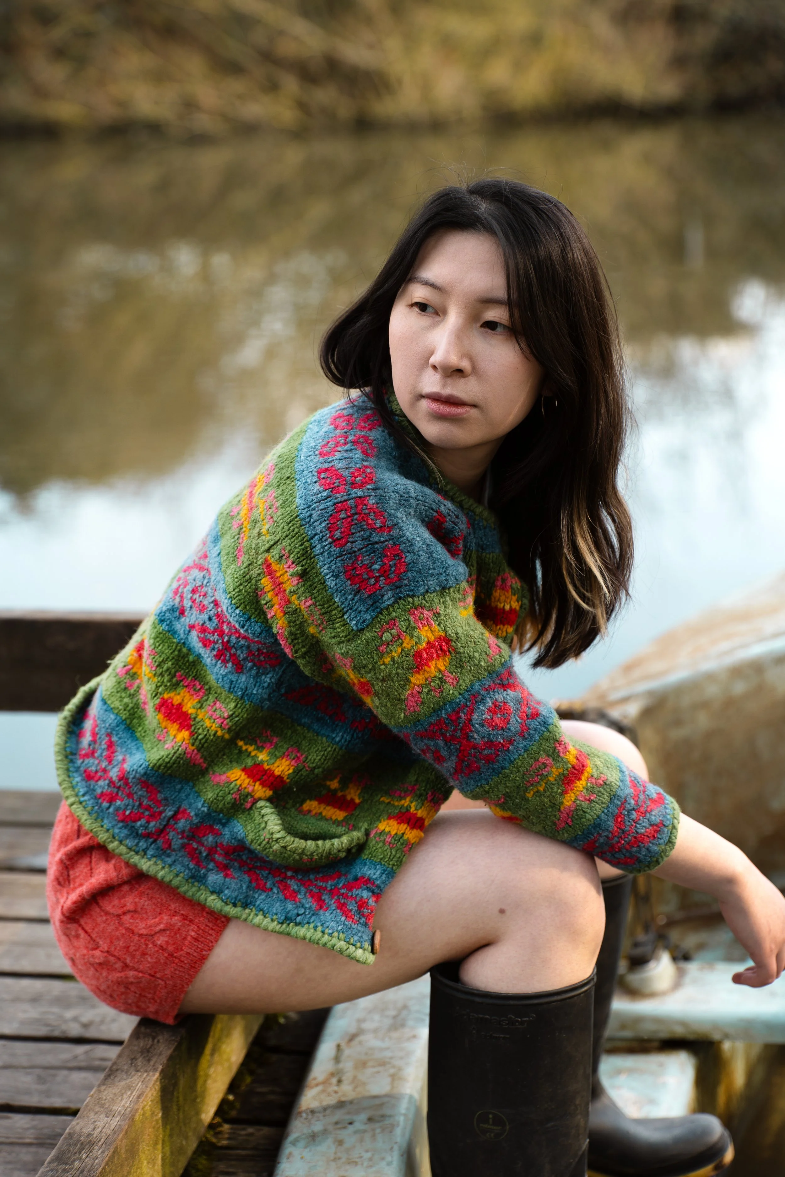A woman with long dark hair sitting on a wooden dock by a lake, wearing a colorful patterned sweater and black rain boots, looking to the side.