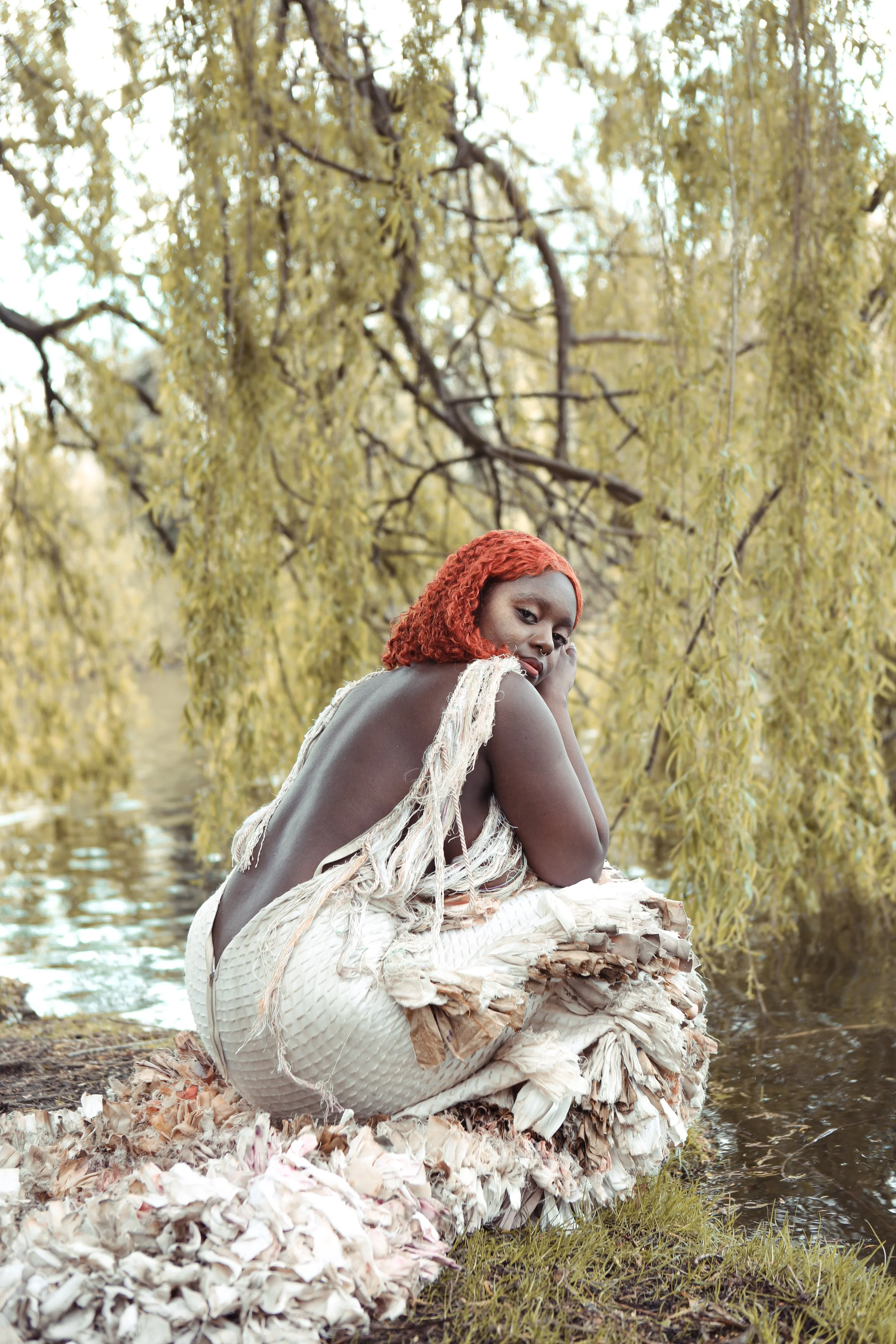 A woman with curly red hair sitting on a piece of driftwood near water, surrounded by yellowish weeping willow trees, wearing a textured, light-colored garment with frayed edges.