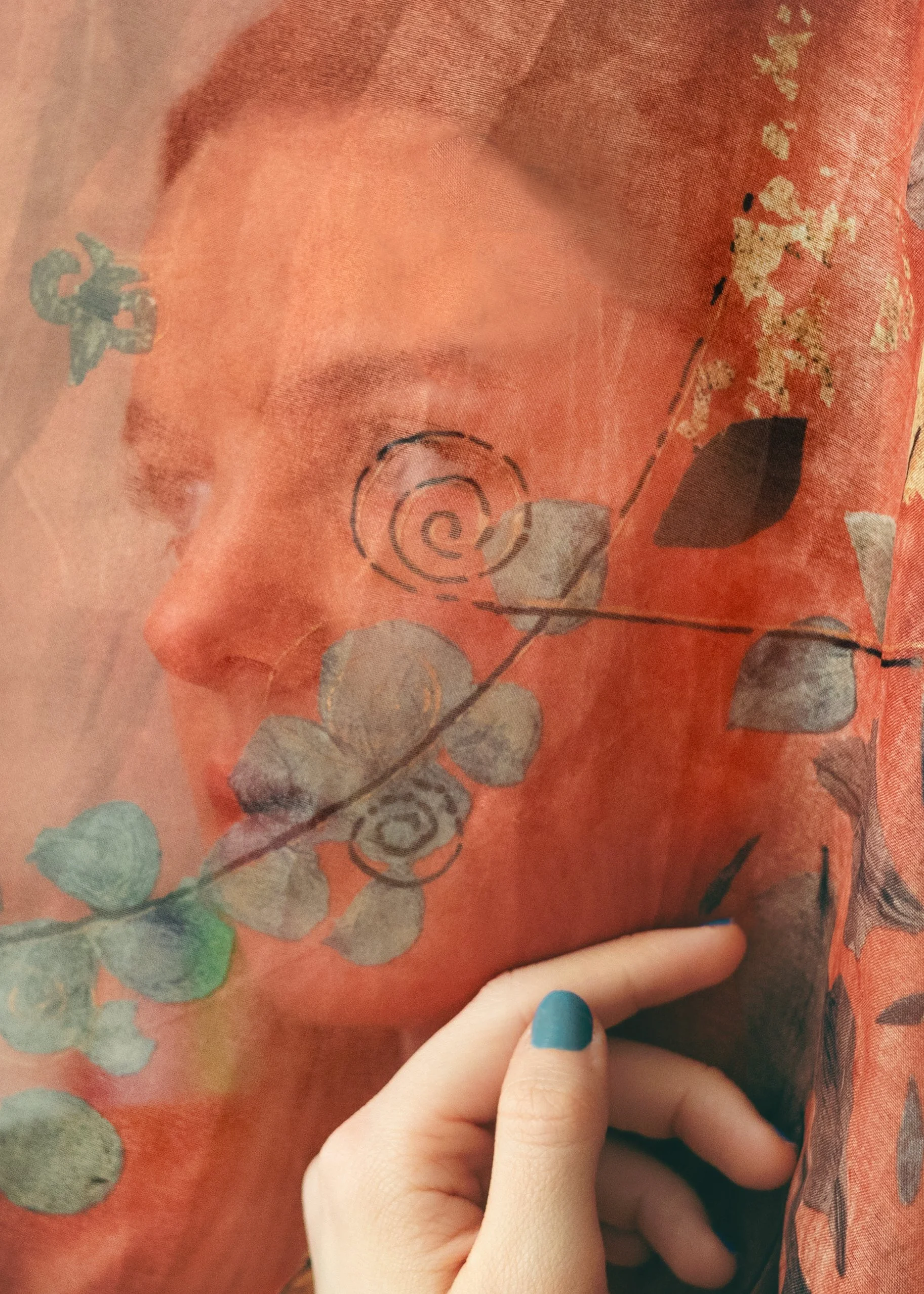 A woman's face is visible behind a semi-transparent floral fabric with a person’s hand gently touching the fabric.