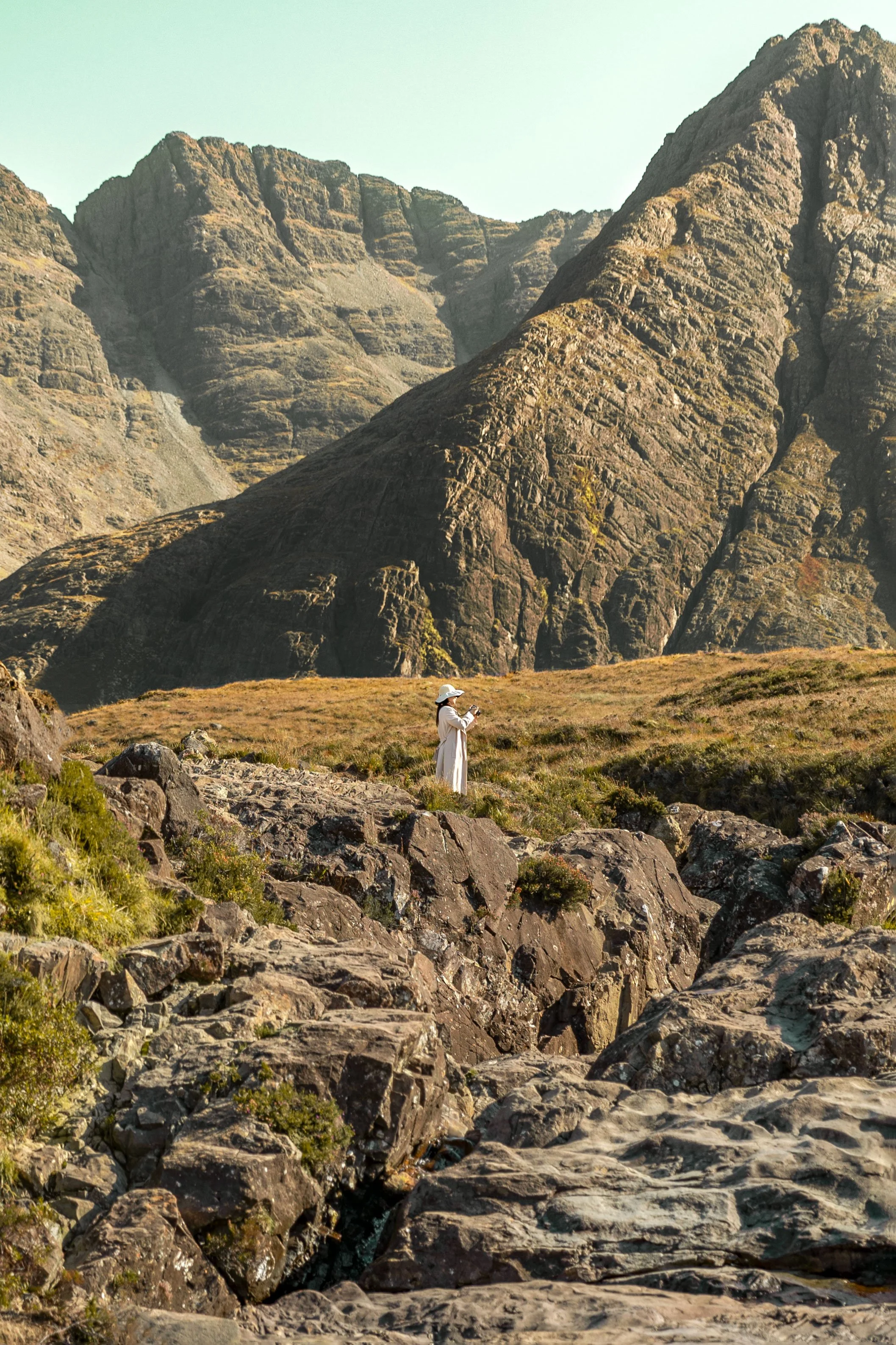 A person in a white dress and hat taking a photo with a phone in rocky, mountainous terrain.