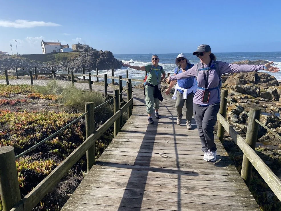 Group of four women walking on a wooden coastal boardwalk with ocean and rocky shoreline in the background, two women extending their arms outward.