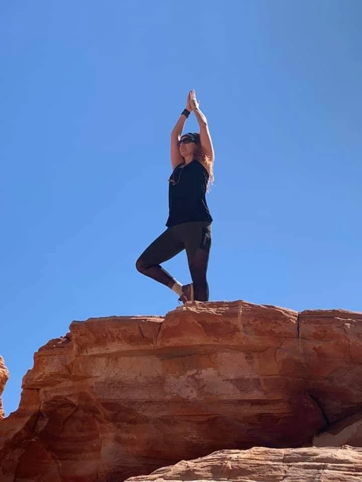 Woman practicing yoga in a tree pose standing on a rock formation with a clear blue sky background.