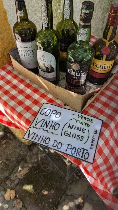 A cardboard box containing six bottles of Portuguese wine on a red and white checkered tablecloth. In front of the box is a handwritten sign listing wine-related items in Portuguese, such as "copo" (glass), "vinho" (wine), "vinho do porto" (port wine
