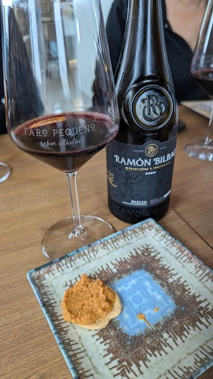 A glass of red wine, a bottle of Ramón Bilbao red wine, and a small serving of food on a decorative plate on a wooden table.