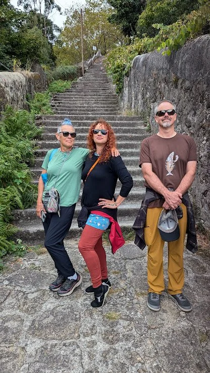 Three people standing on stone steps outdoors, with greenery and trees around.