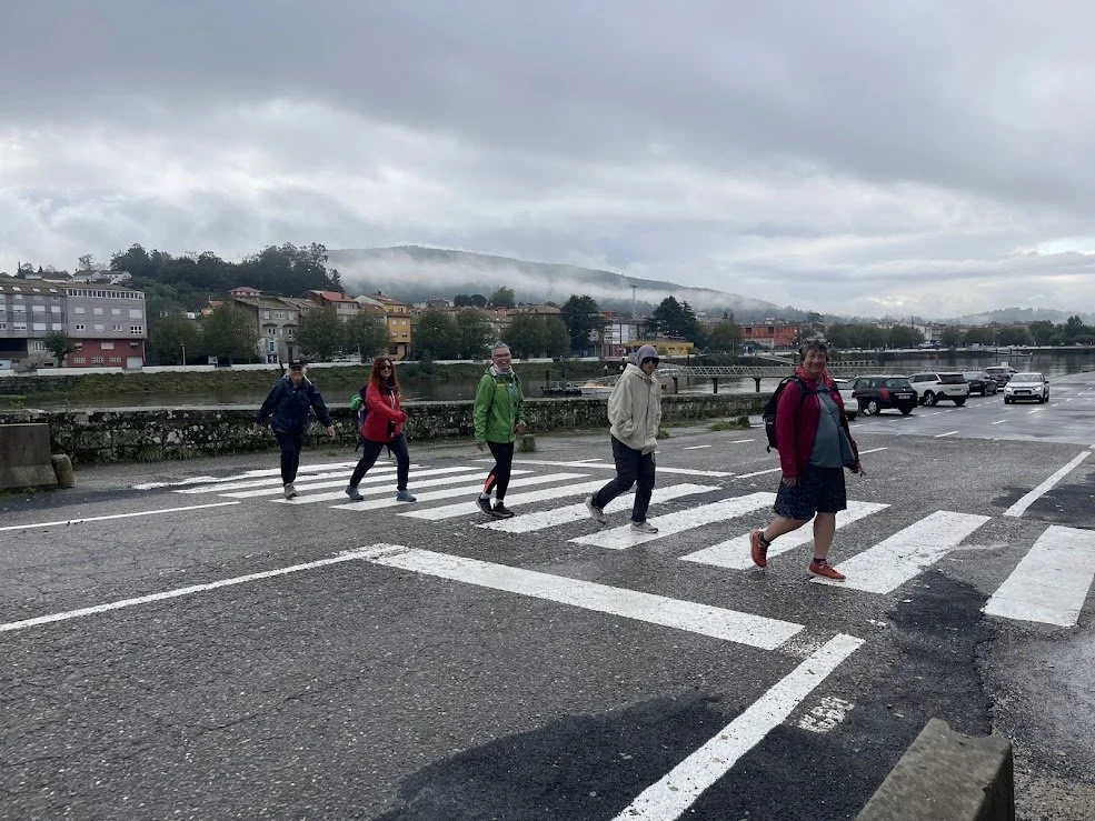 Five people walking on a crosswalk near a river, with a town and hills in the background on a cloudy day.