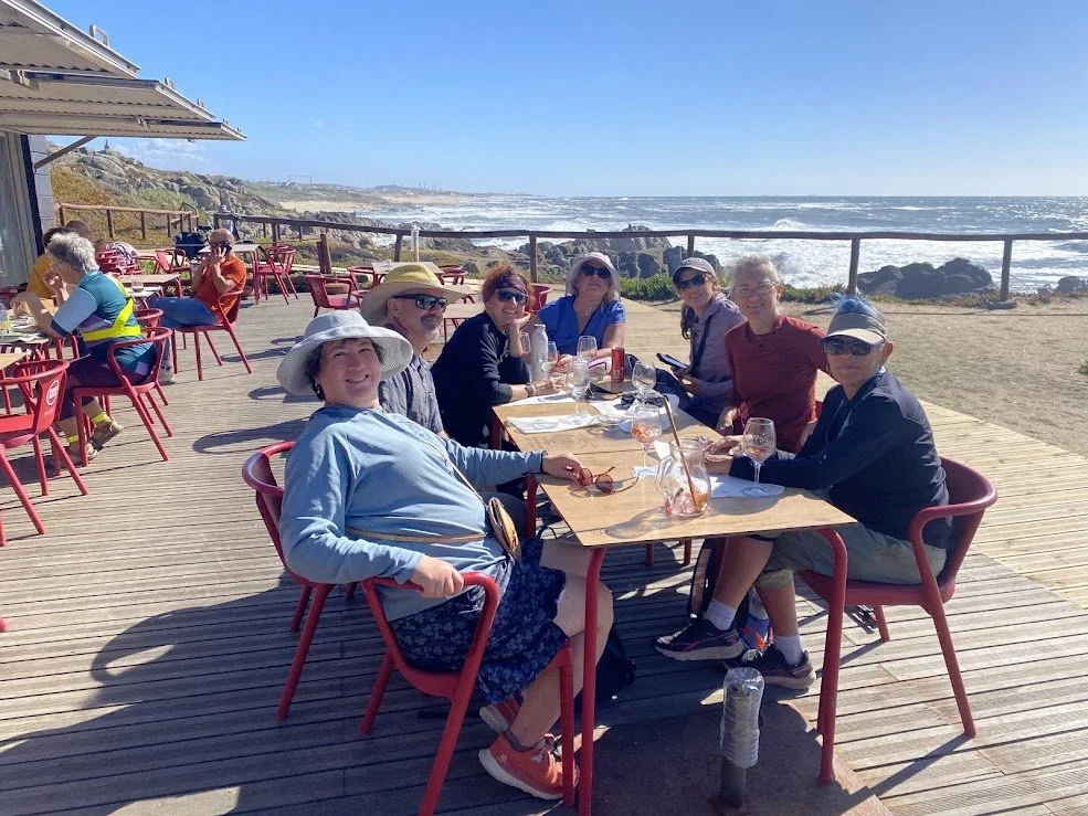 A group of seven women enjoying a meal at a seaside restaurant with a view of the ocean and rocks in the background on a sunny day.