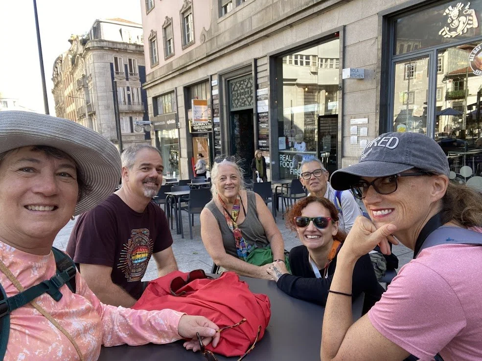 Six people sitting around an outdoor table on a city street, smiling and enjoying a sunny day. The group includes three women wearing sunglasses and two men, with various buildings and shop windows visible in the background.