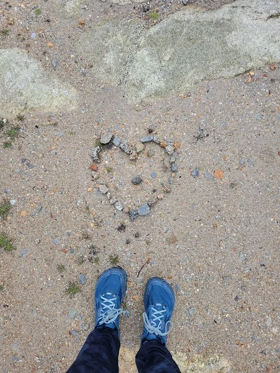 A person's feet in blue shoes standing on sandy ground near a rock formation and a heart shape made of small rocks.