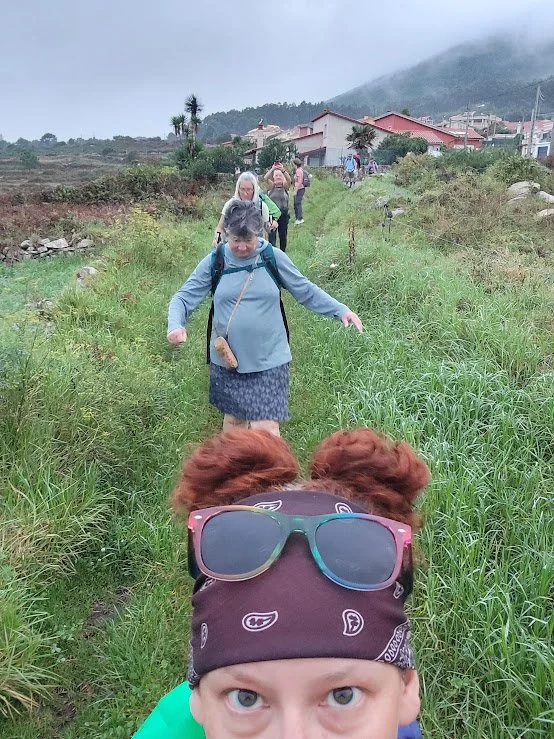 Child with red hair, wearing a bandana and sunglasses on head, takes a selfie on a grassy trail with a group of people walking ahead in a rural, mountainous area.