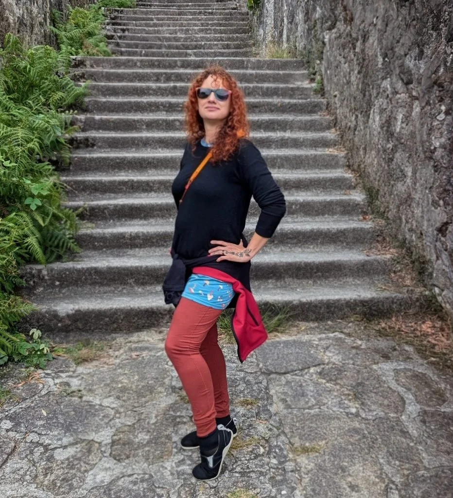 A woman with curly red hair wearing sunglasses, a black long-sleeve shirt, brown pants, and black shoes, standing outdoors on stone steps beside a stone wall, with green plants on the side.