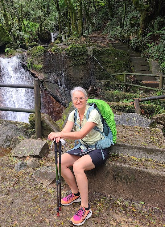 A woman with gray hair, glasses, and a bright smile sitting on a rock in a lush, green forest near a waterfall, carrying a large green backpack and holding trekking poles.