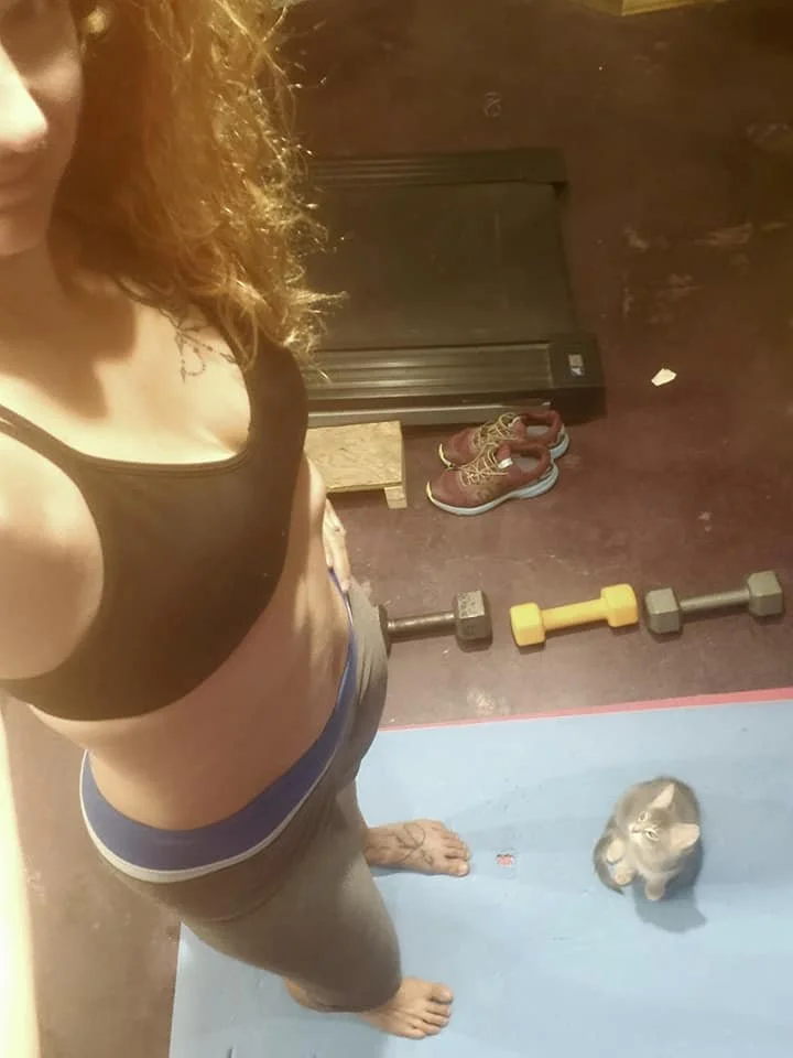 Top-down view of a person wearing a black sports bra and gray shorts standing on a blue exercise mat in a gym. There is a small gray and white cat on the mat, facing the camera. Gym equipment, including a yellow dumbbell, a gray dumbbell, and a barbell, are on the floor nearby. Shoes are on a small wooden shelf against the wall.