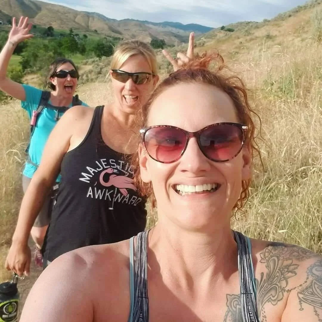 Three women taking a selfie on a trail in a hilly outdoor area with grassy fields and mountains in the background, smiling and making peace signs.