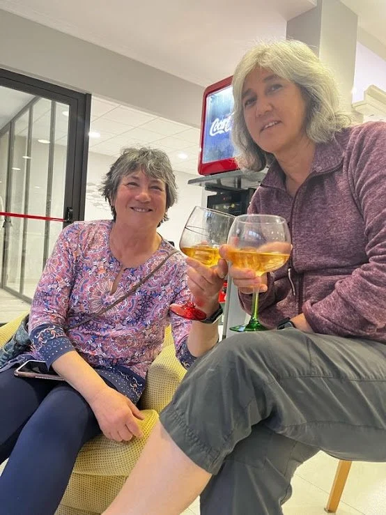 Two women sitting indoors, smiling, holding glasses of a golden beverage, possibly wine or a cocktail, with a Coca-Cola vending machine in the background.