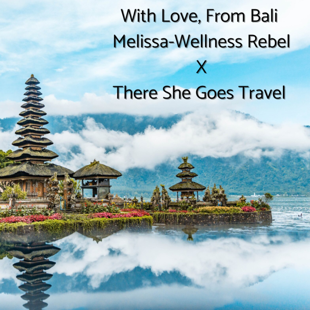 Scenic view of traditional Balinese temple structures with tiered roofs, colorful flowers, and a reflecting pool, with mountains and clouds in the background.