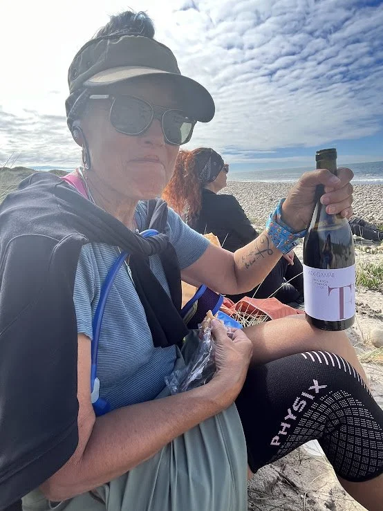 A woman sitting on a beach holding a bottle of wine and a plastic cup in her hand, wearing sunglasses, a cap, and a backpack, with another person sitting behind her, under a partly cloudy sky.