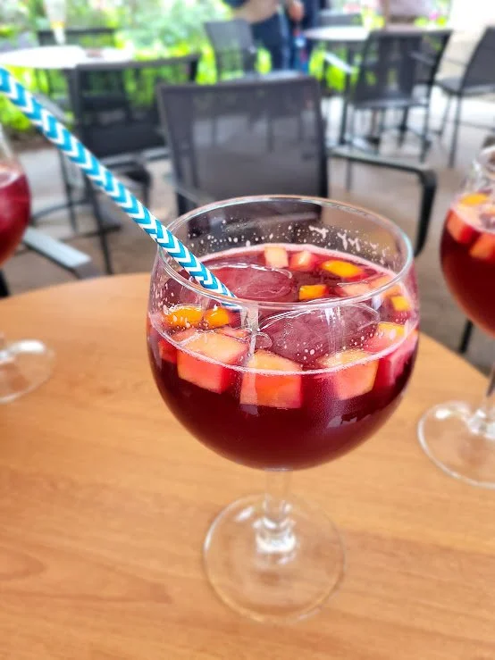 A glass of red sangria with fruit pieces and a blue striped straw on a wooden table. In the background, there are outdoor chairs and a blurred patio setting.