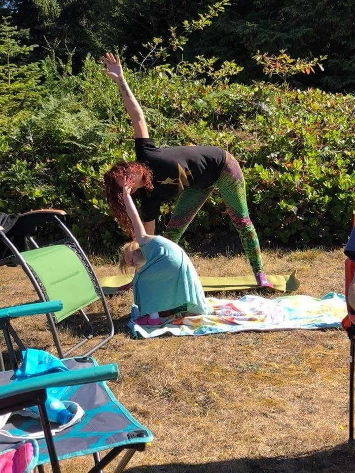A woman and a young girl practicing yoga outdoors on a sunny day. The woman is in a bent-over yoga pose, reaching down to the girl who is on her knees, also reaching up. They are on mats on the grass, surrounded by outdoor chairs and greenery.