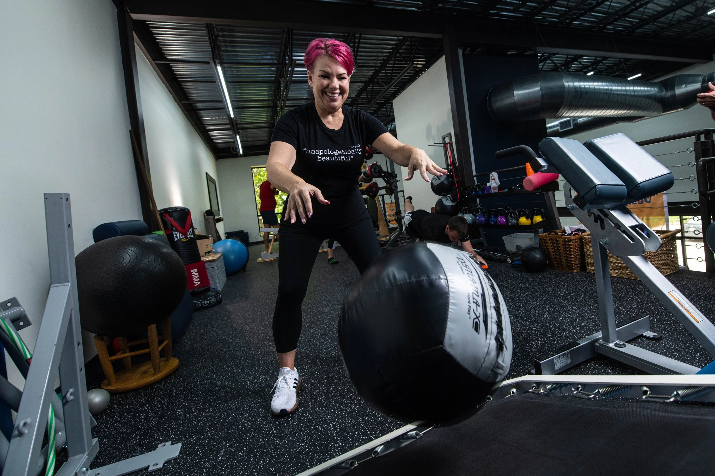 Woman with pink hair smiling in a gym, preparing to roll a large black and white punching bag on the floor. Gym equipment and other people working out are visible in the background.