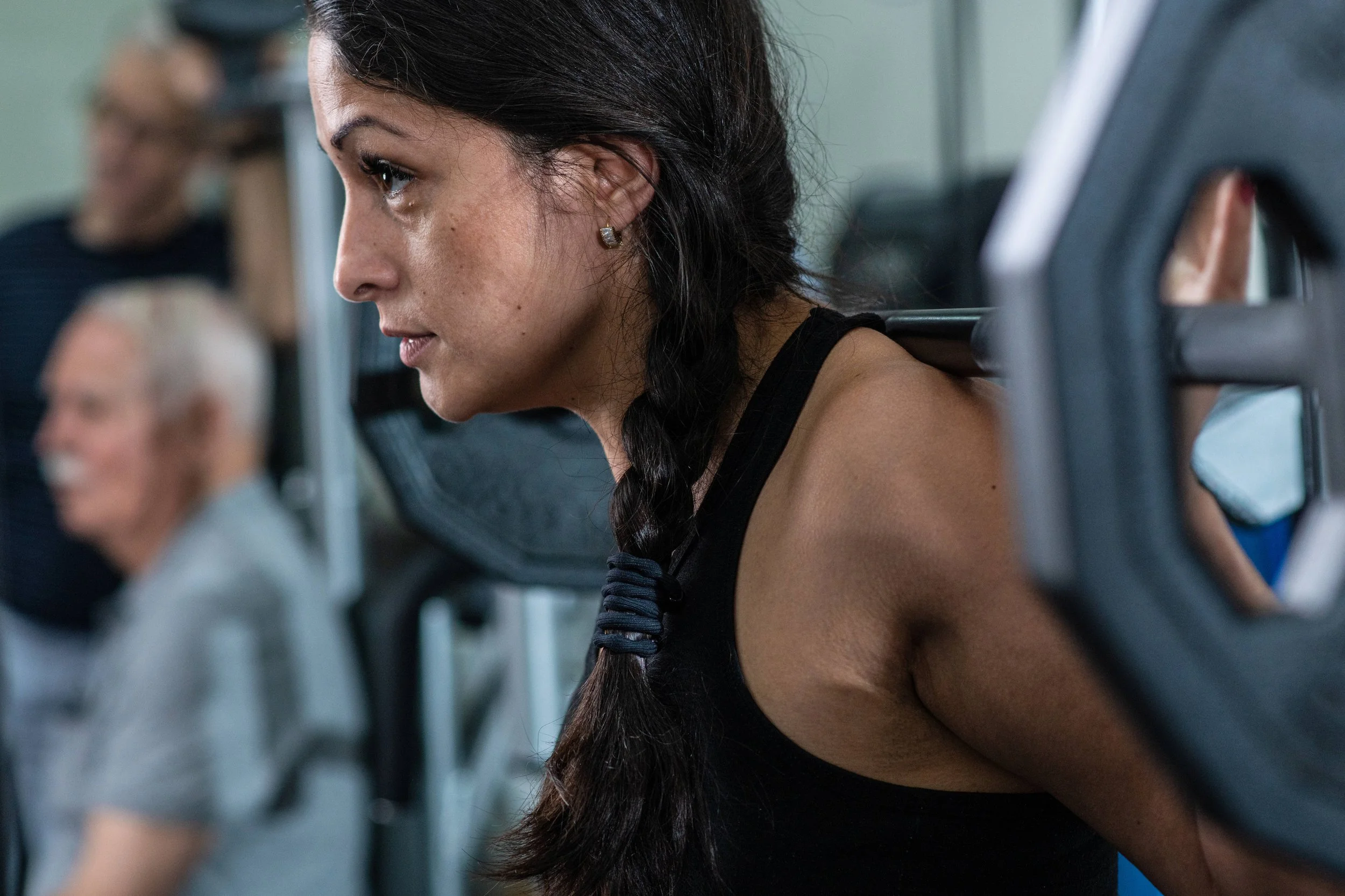 A woman with long dark hair, wearing a black workout top, is sitting on a bench in a gym, focusing on her workout. There are other people exercising in the background.