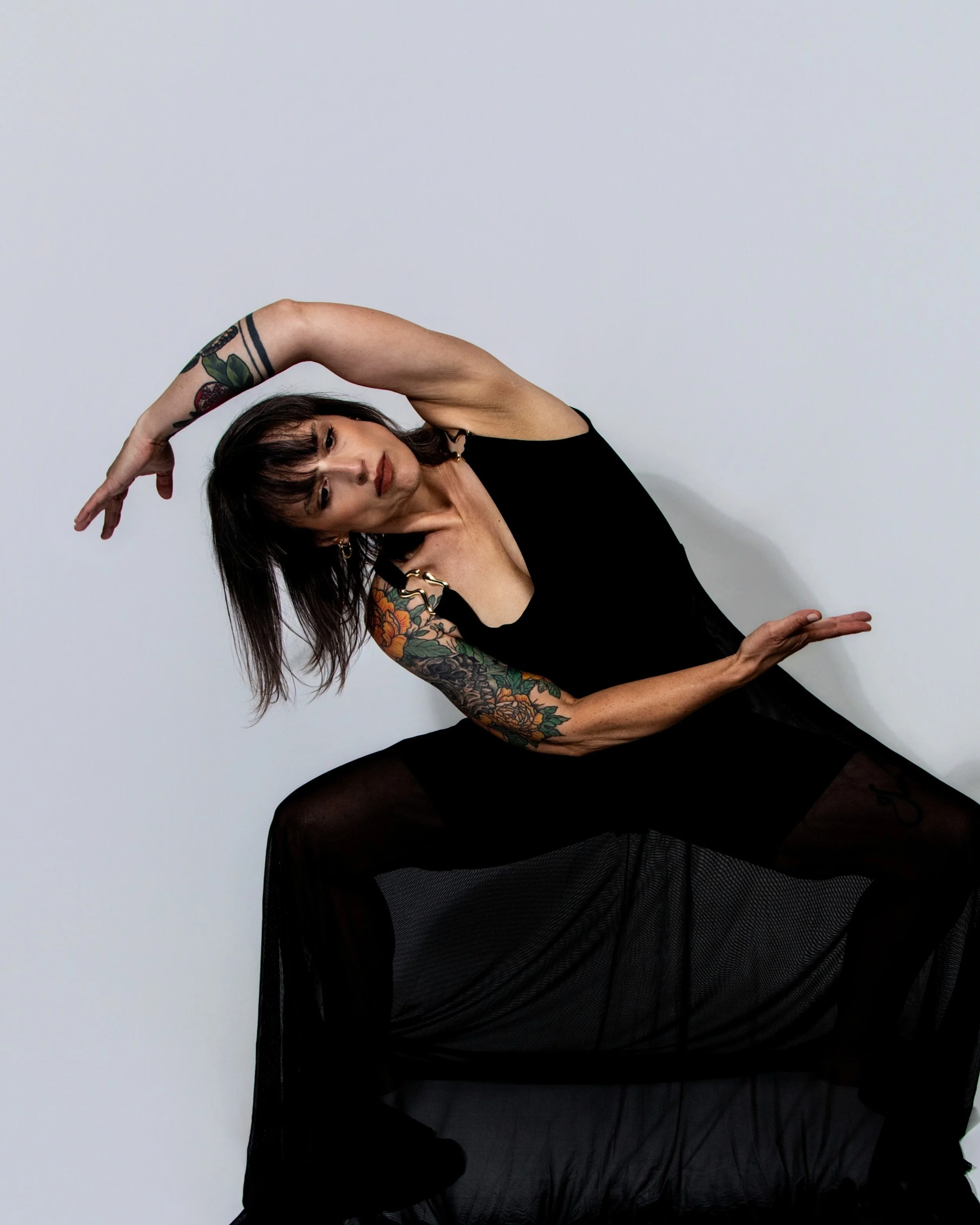 A woman with short dark hair and tattoos performing a dance pose against a plain white wall, wearing a black dress and black tights.
