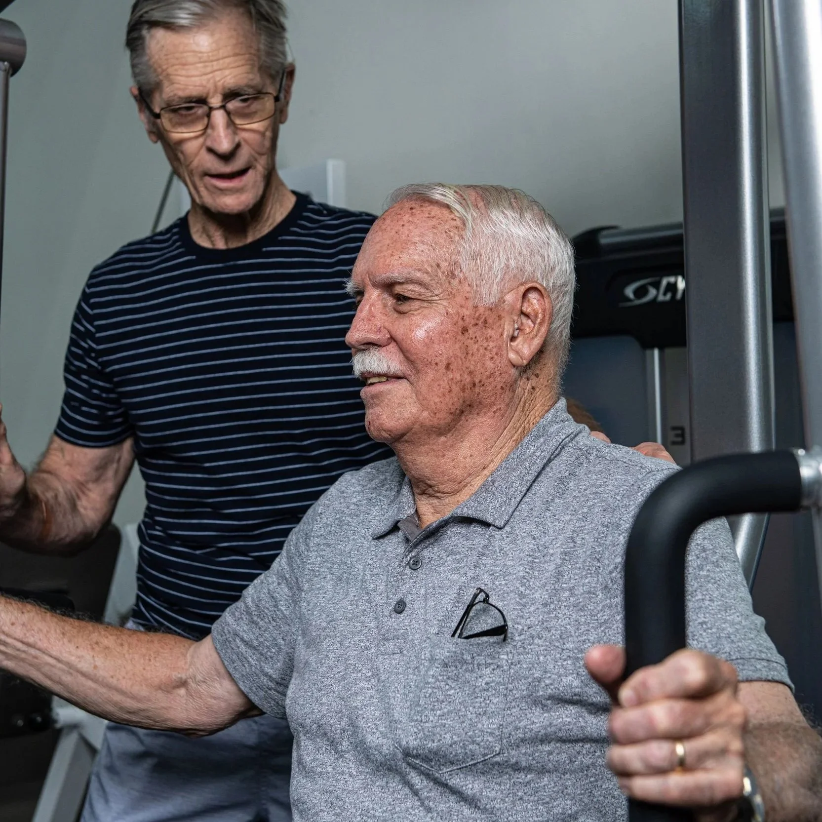 An elderly man with some sun spots on his face, wearing a gray polo shirt, is seated in a wheelchair, smiling, with a man's arm around his shoulder. Another older man with glasses, wearing a striped shirt, is standing next to him, slightly leaning in, and appears to be talking to him.