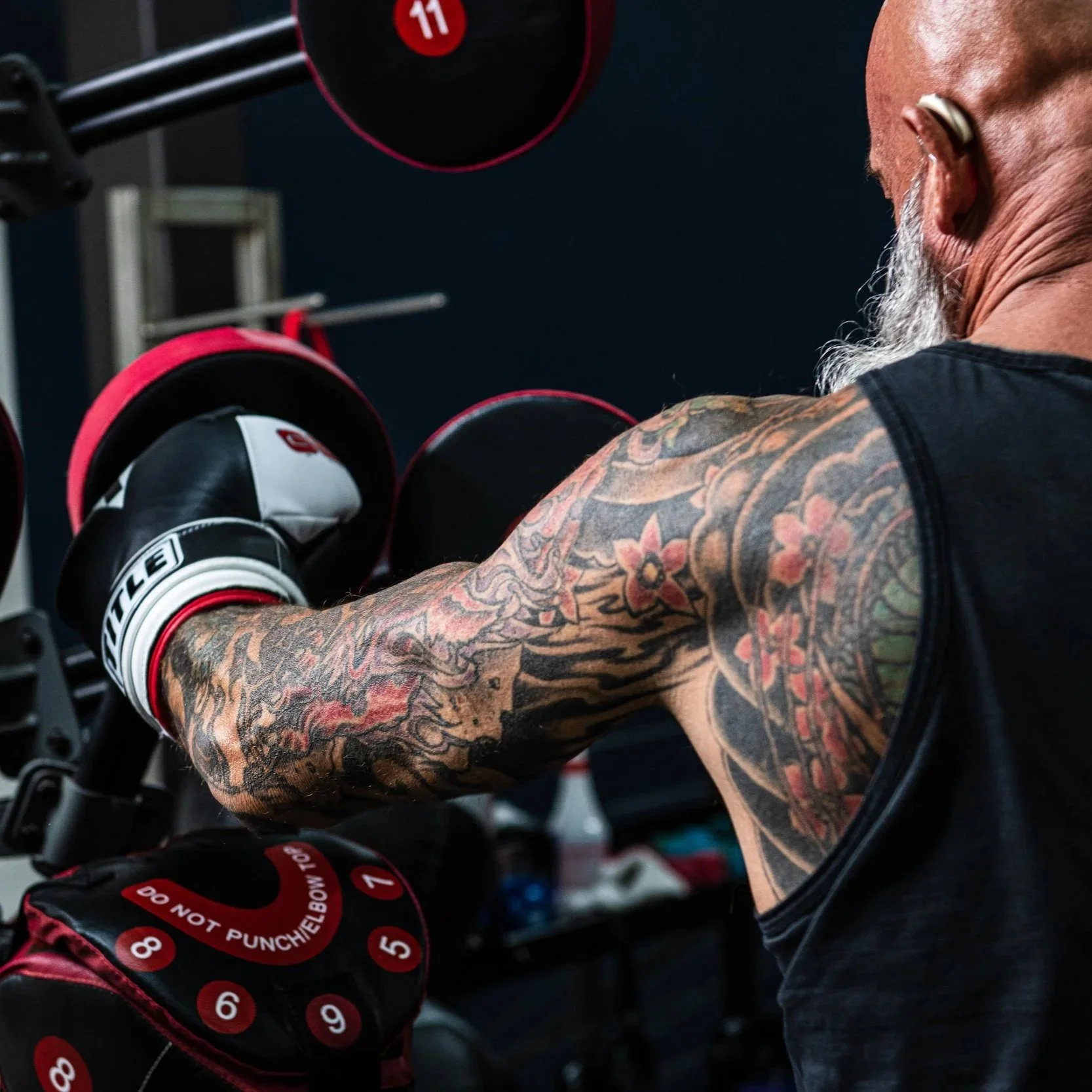 A person with a heavily tattooed right arm wearing black sleeveless shirt and white boxing gloves with red accents, lifting a dumbbell in a gym.
