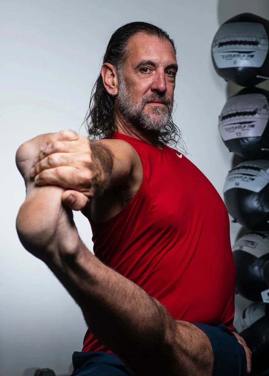 A middle-aged man with long dark hair and a beard, wearing a red workout shirt, stretching his arm in a gym or sports facility.