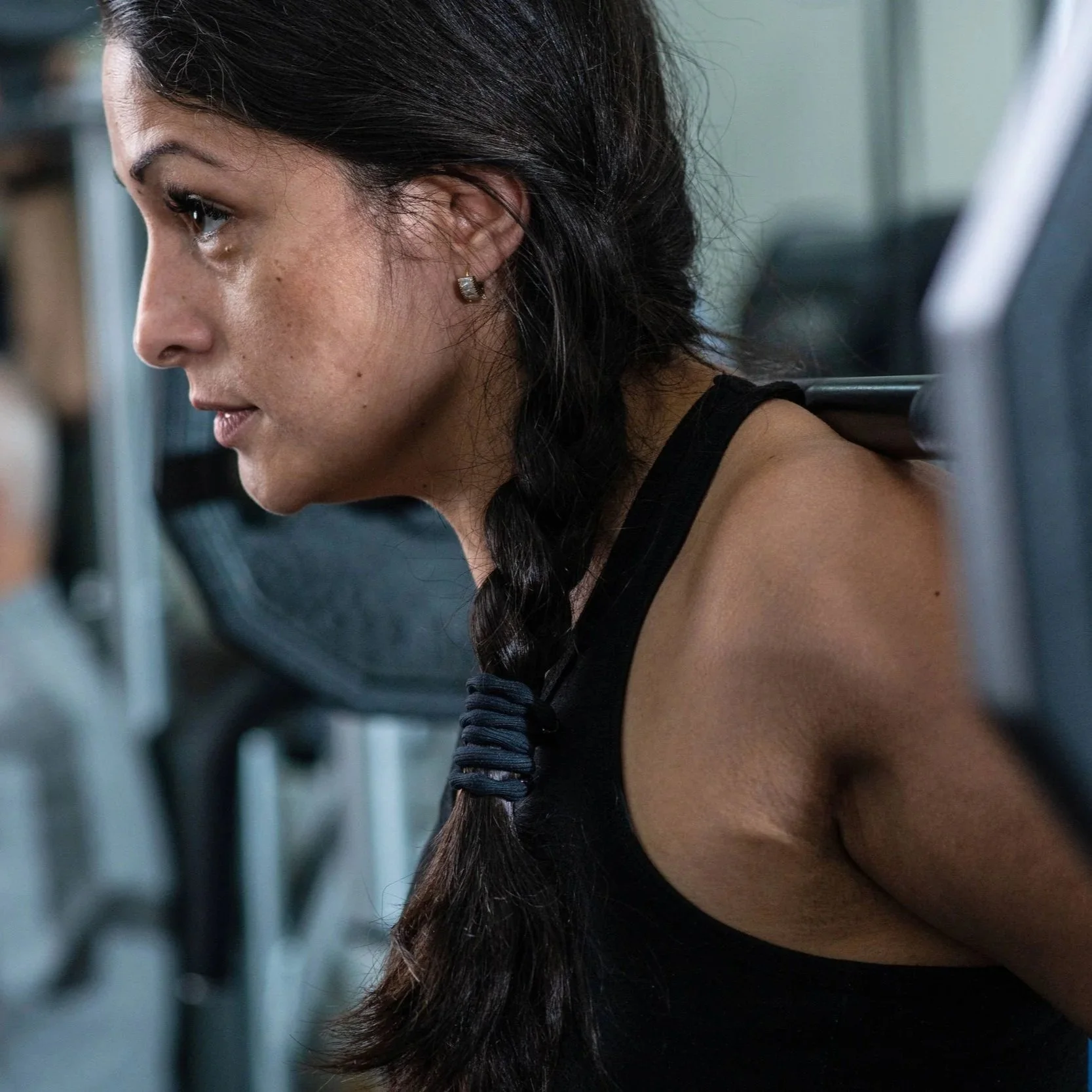 Close-up side view of a woman with long dark hair in a braid, wearing diamond stud earrings, in a gym or fitness environment.