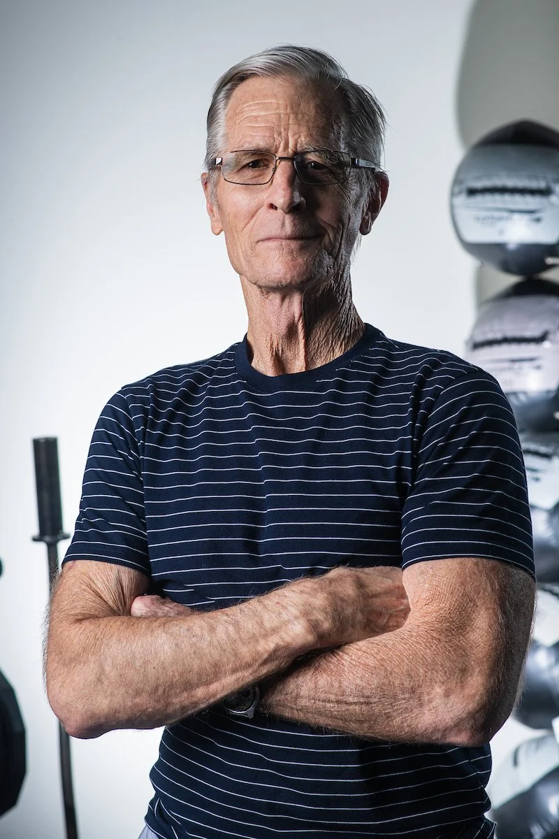 An older man with gray hair and glasses, wearing a navy blue striped t-shirt, standing with arms crossed, in a room with a white wall and a shelf with sports helmets or similar objects.
