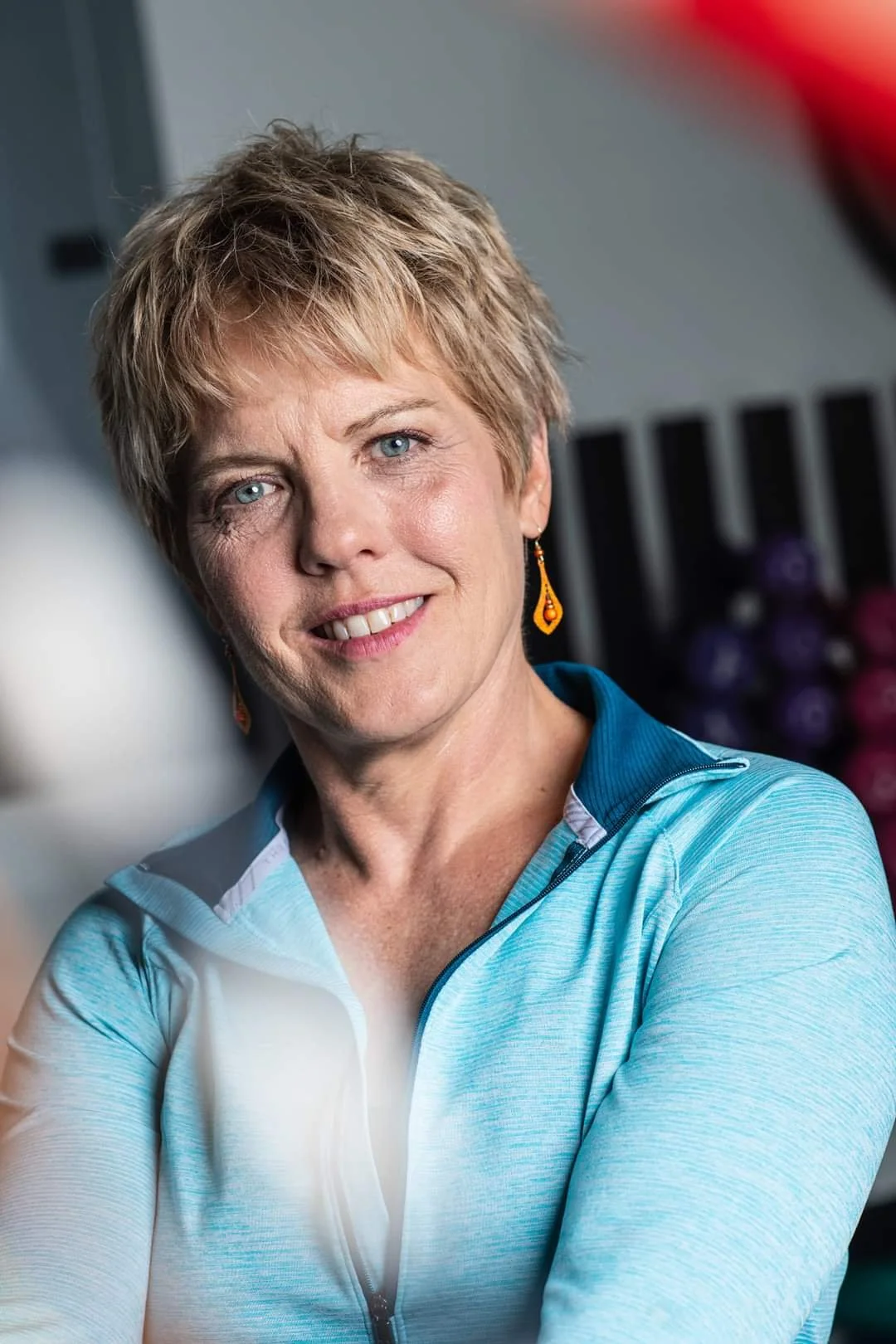 A middle-aged woman with short blonde hair wearing a blue sports jacket and orange earrings, smiling in a gym with weights in the background.