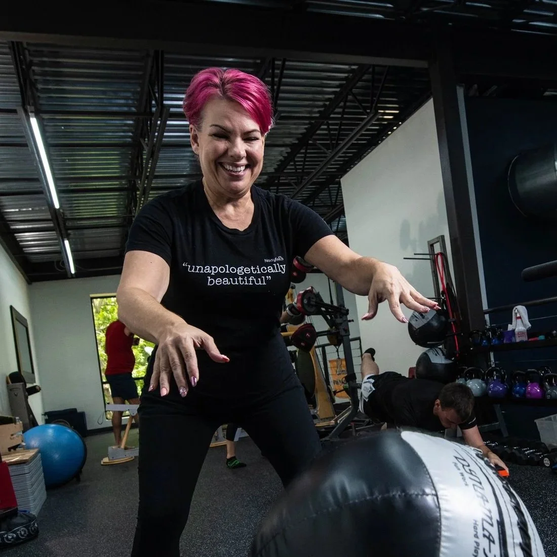 Woman with bright pink hair smiling and playing with a medicine ball in a gym, with other people exercising in the background.
