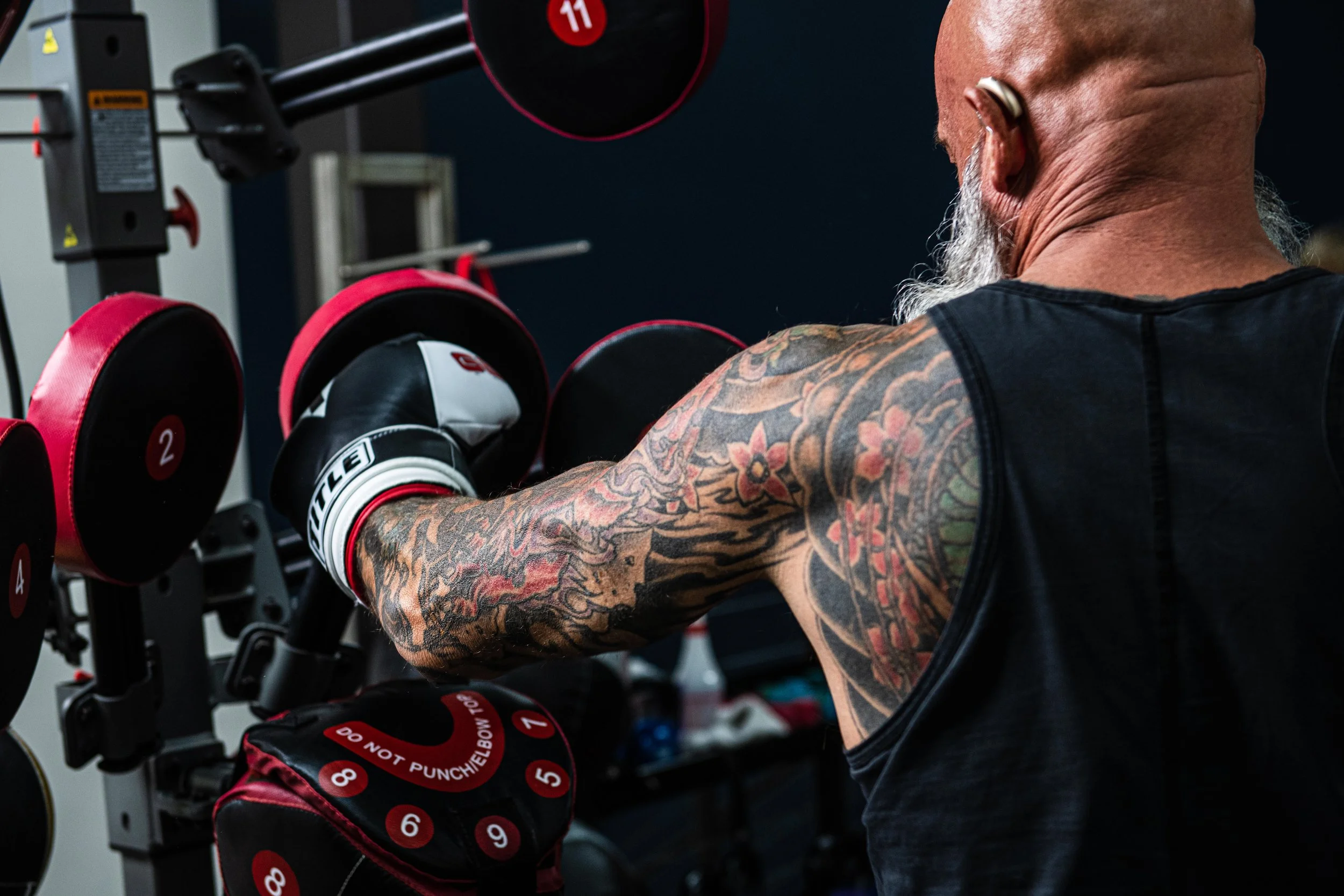 A man with a tattooed arm working out with dumbbells in a gym, wearing boxing gloves and a black sleeveless shirt. There are various weight plates in the background.
