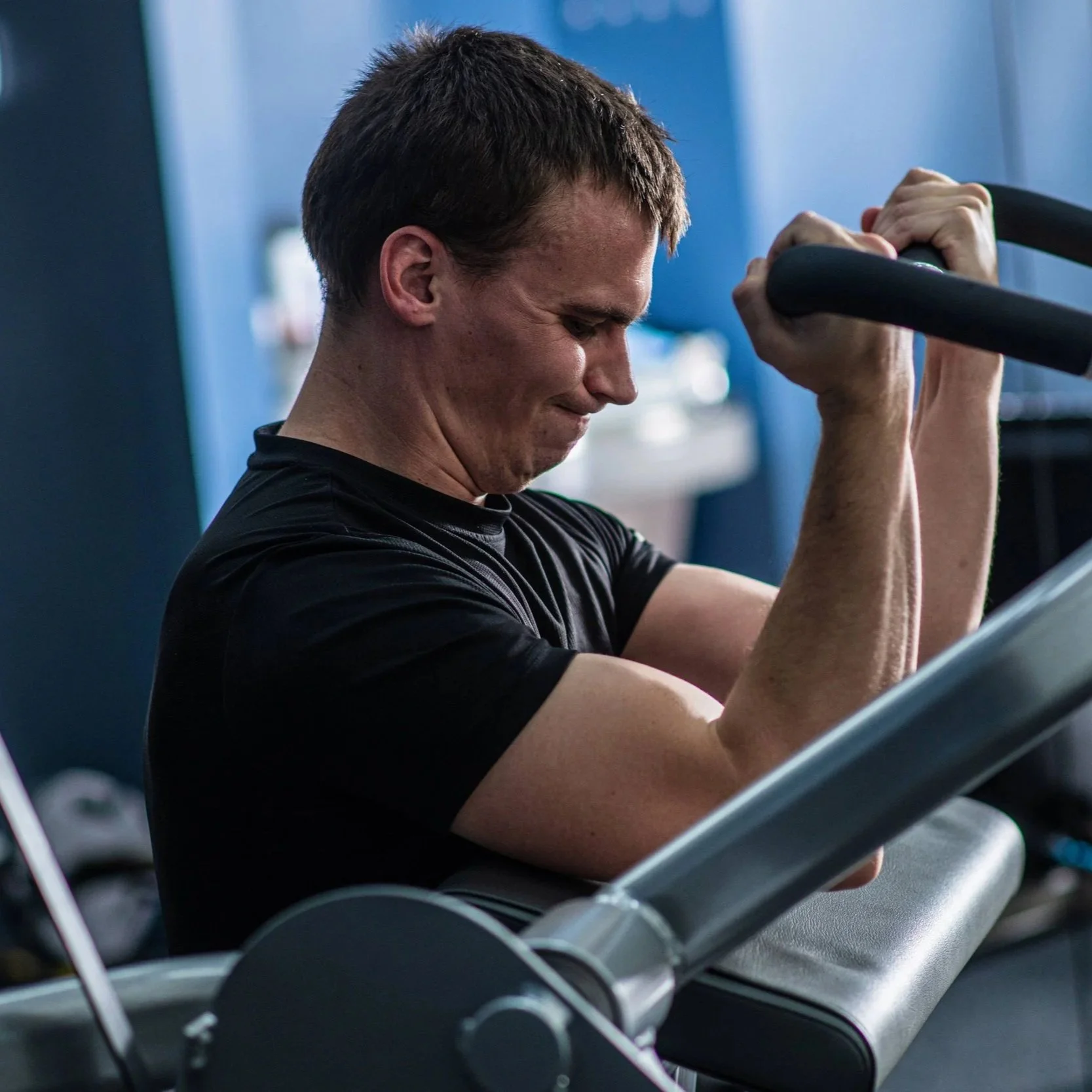 A man with short brown hair, wearing a black shirt, is exercising using an exercise machine in a gym. He is smiling and appears to be focused on his workout.