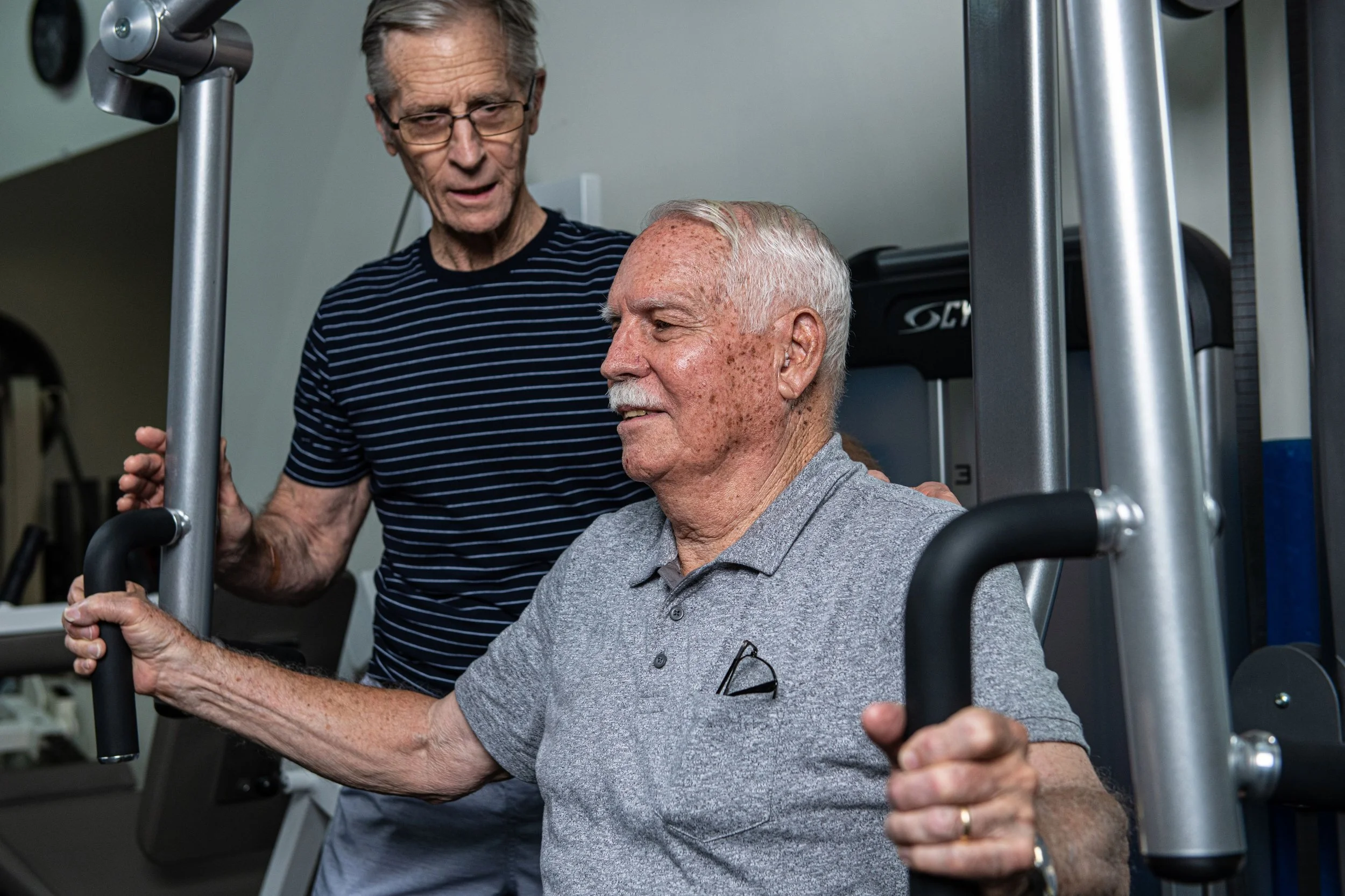 An elderly man performing a seated shoulder press exercise with a trainer assisting him in a gym.