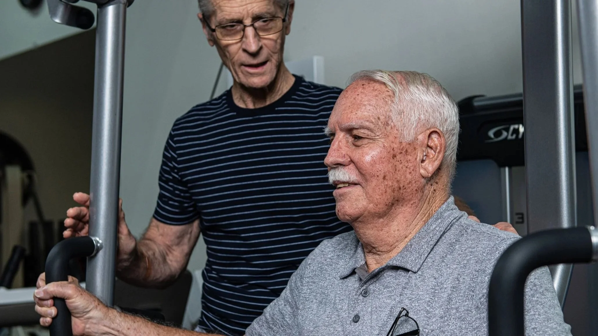 An elderly man with gray hair and freckles doing a seated exercise at the gym while a trainer stands behind him, offering encouragement.