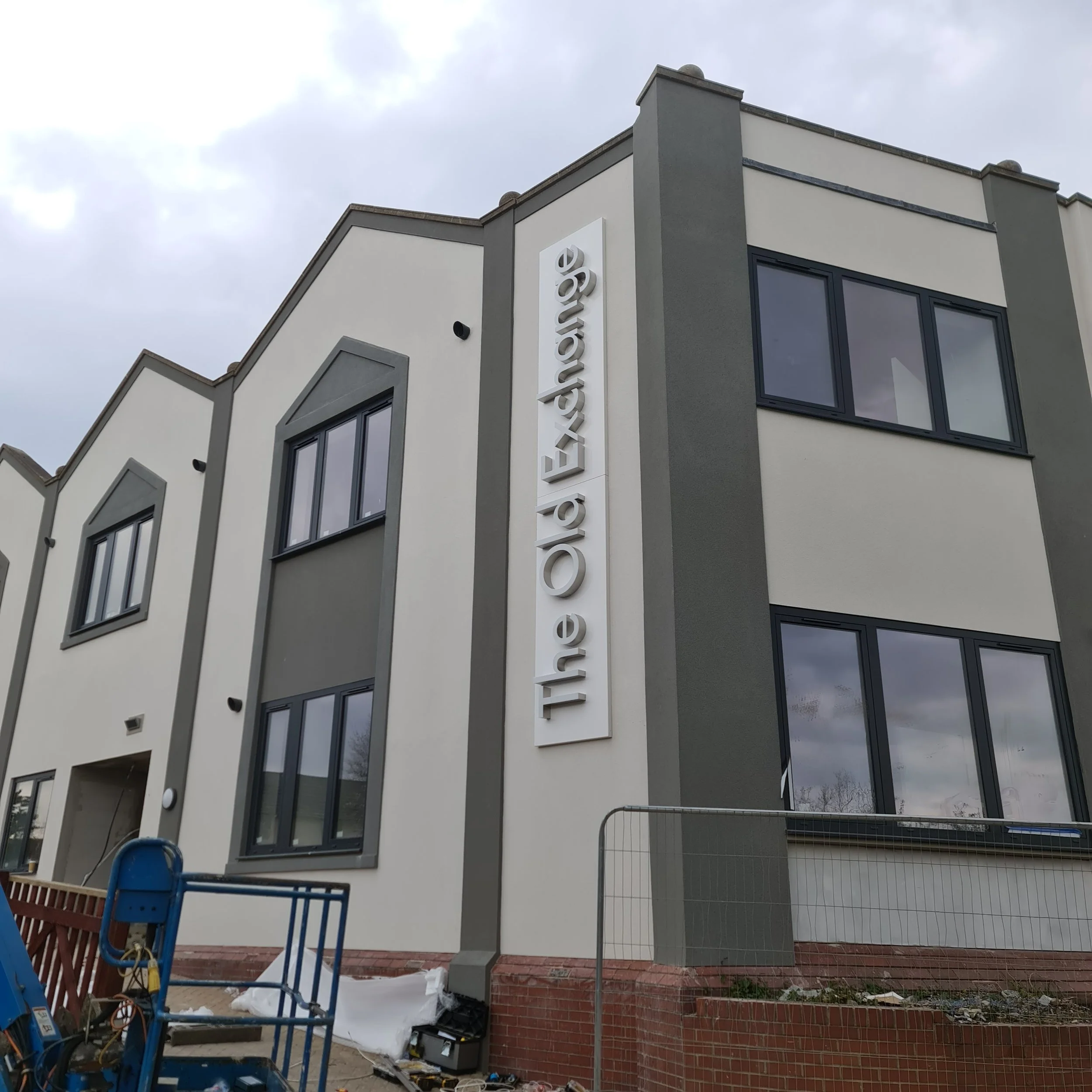 Construction site with a modern multi-story residential building featuring gray and white exterior walls, large black-framed windows, and a vertical sign that reads 'excellence' on the facade. There is construction equipment, including a blue lift, a
