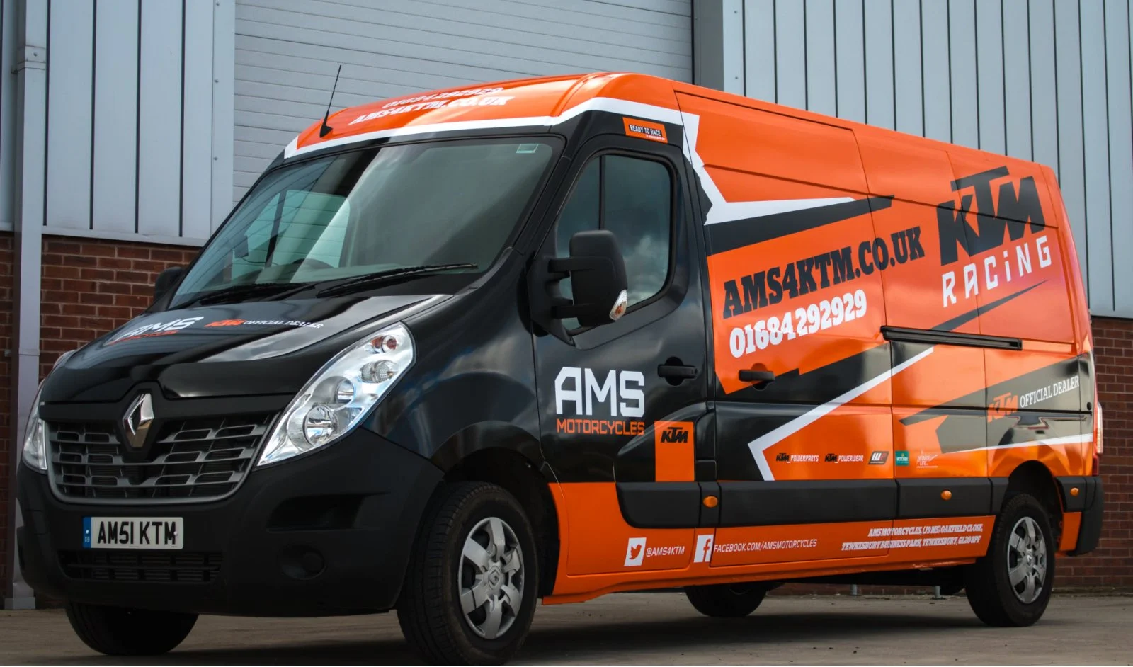 Black and orange racing team van with ' KTM Racing branding and contact information, parked in front of a building.