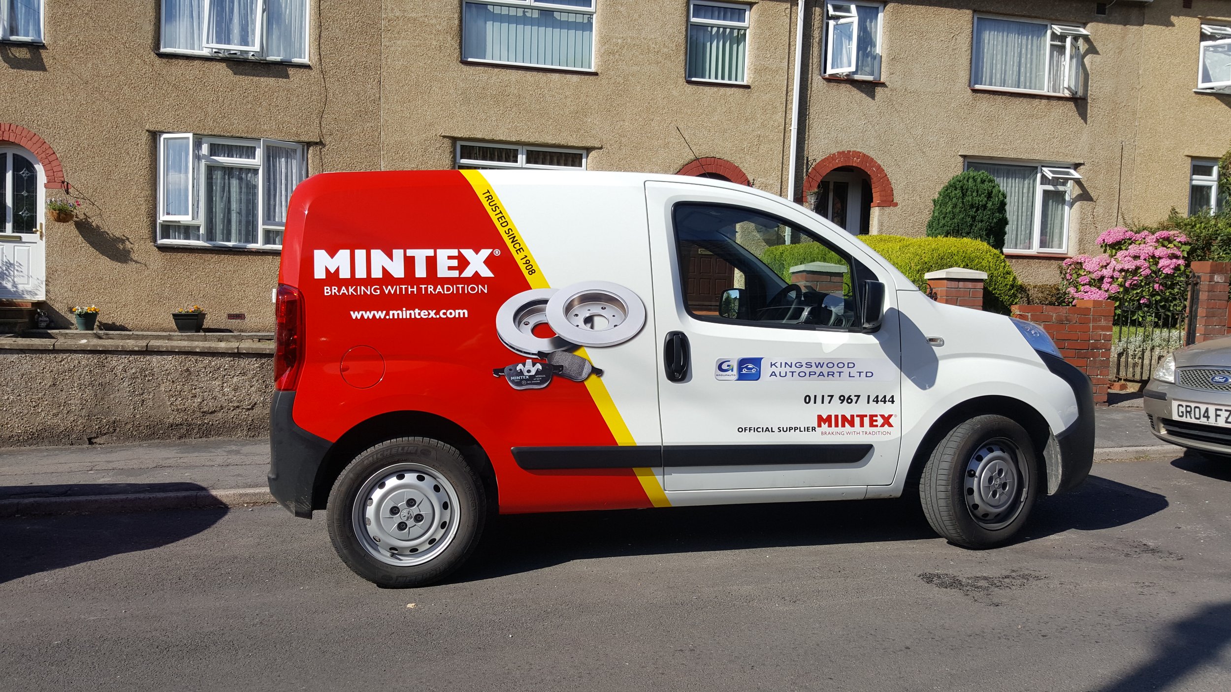 A white commercial van parked on a residential street in front of beige apartment building with flowerpots and bushes. The van has Mintex branding, including red and yellow graphics, and a website URL. The van also displays the Kingswood Autopart Ltd